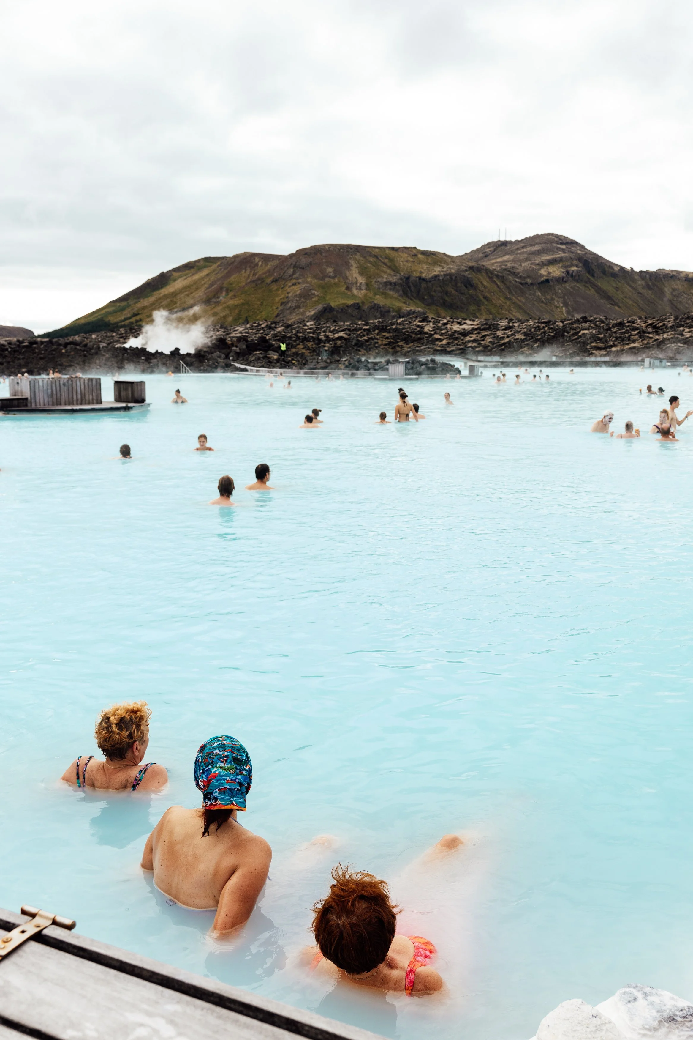 People relaxing and swimming in the blue geothermal waters of the Blue Lagoon, with volcanic mountains in the background, during cloudy weather.
