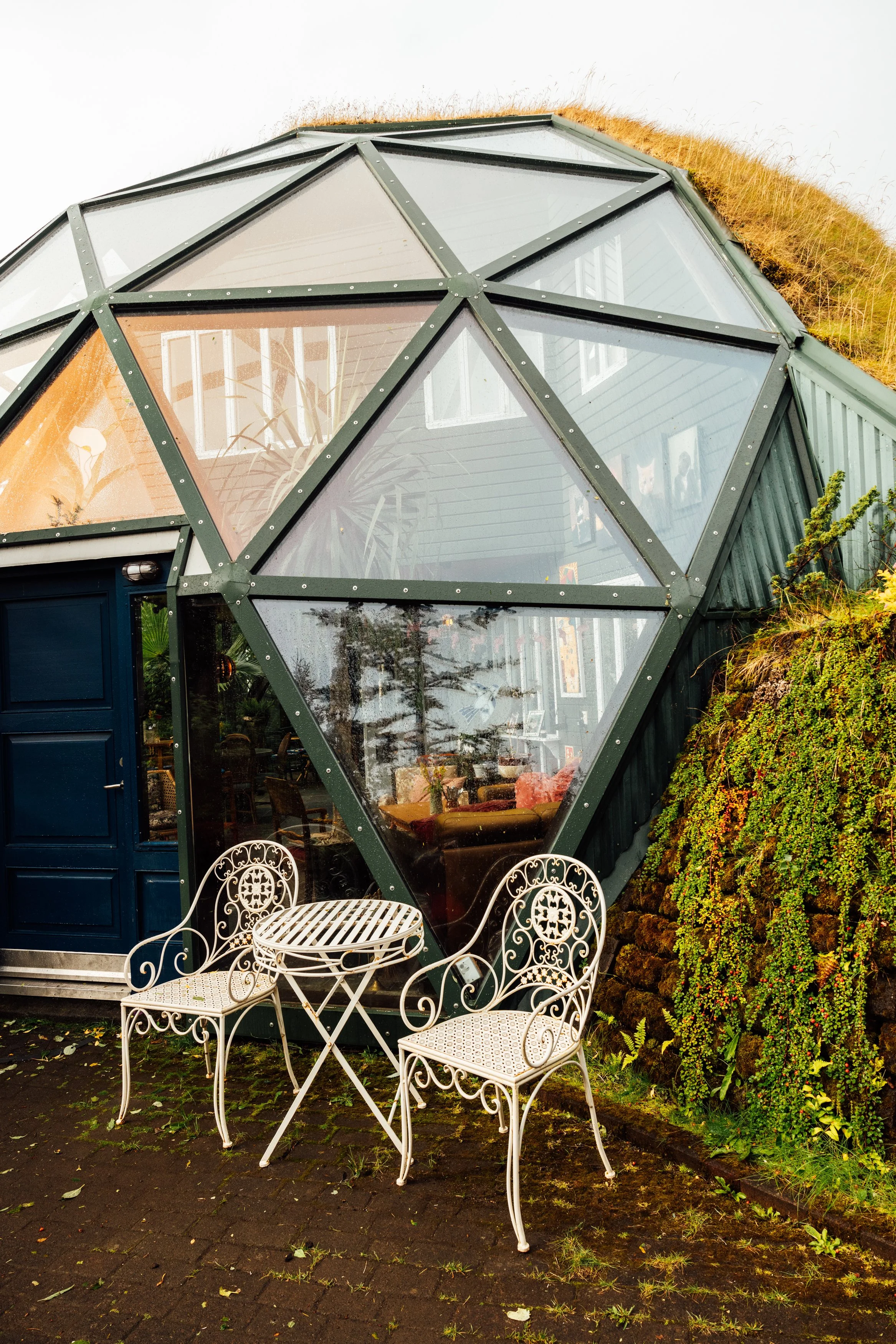 A geodesic dome house with large glass windows, green metal frame, and grass on the roof. Two ornate white metal chairs and a small table sit outside on a brick patio.