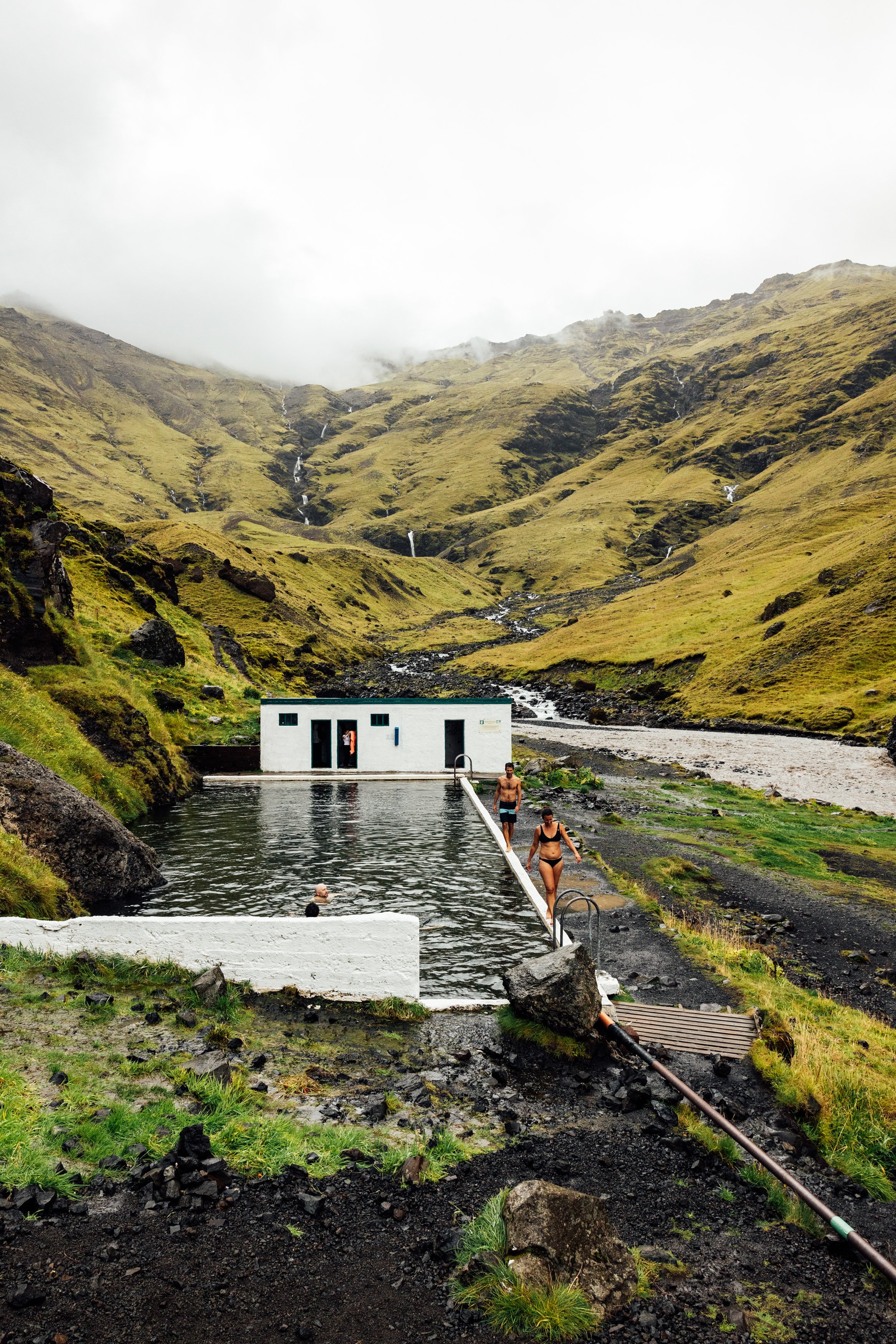 People swimming and walking at a geothermal hot spring pool in a mountain landscape with waterfalls
