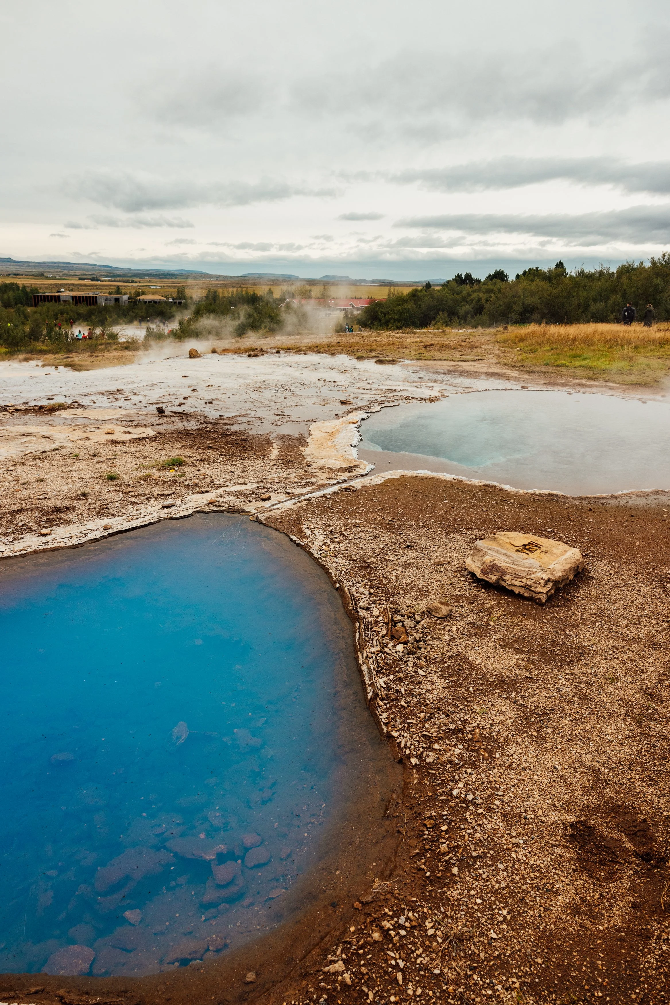 Hot springs with colorful pools, steam rising, in a geothermal landscape with cloudy sky and distant hills.