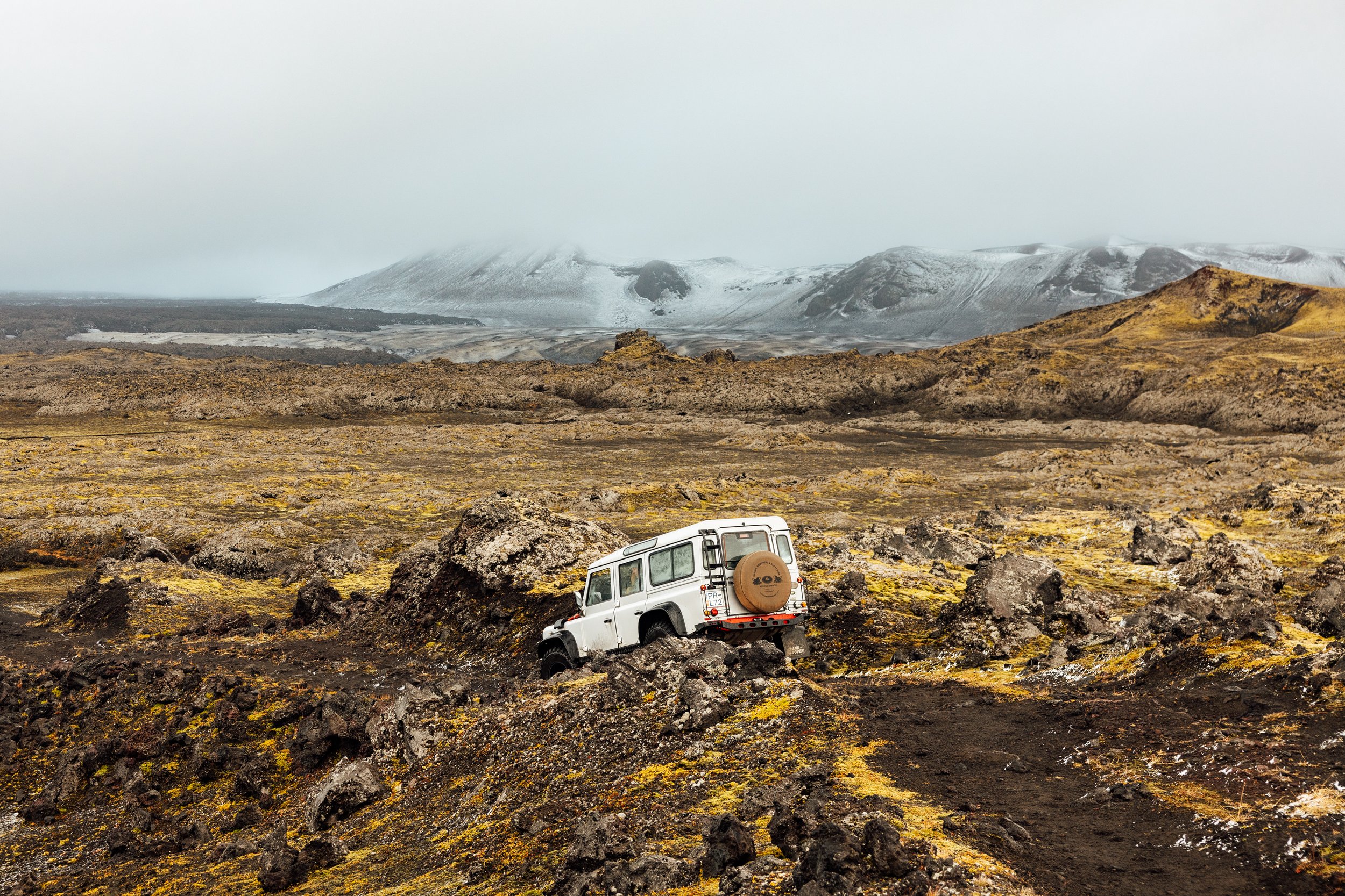 An off-road vehicle navigating a rugged, volcanic landscape during daytime with overcast sky, mountains in the background, and no other vehicles or people visible.