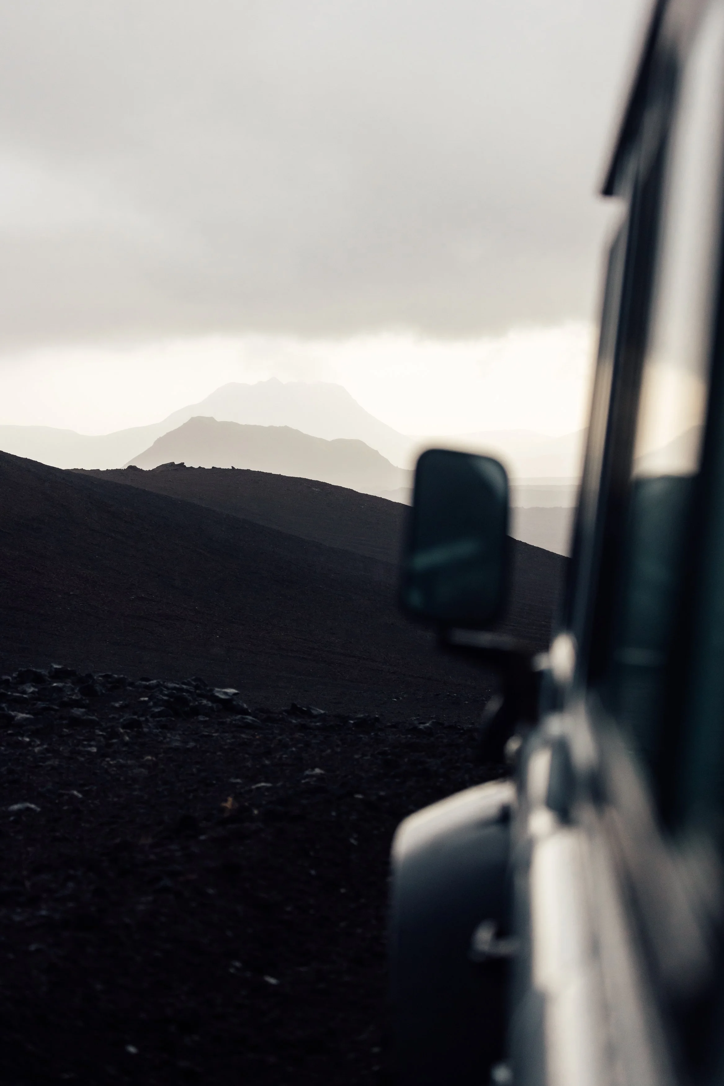 A view from a vehicle showing a rugged landscape with dark, volcanic terrain and distant mountains under a cloudy sky.