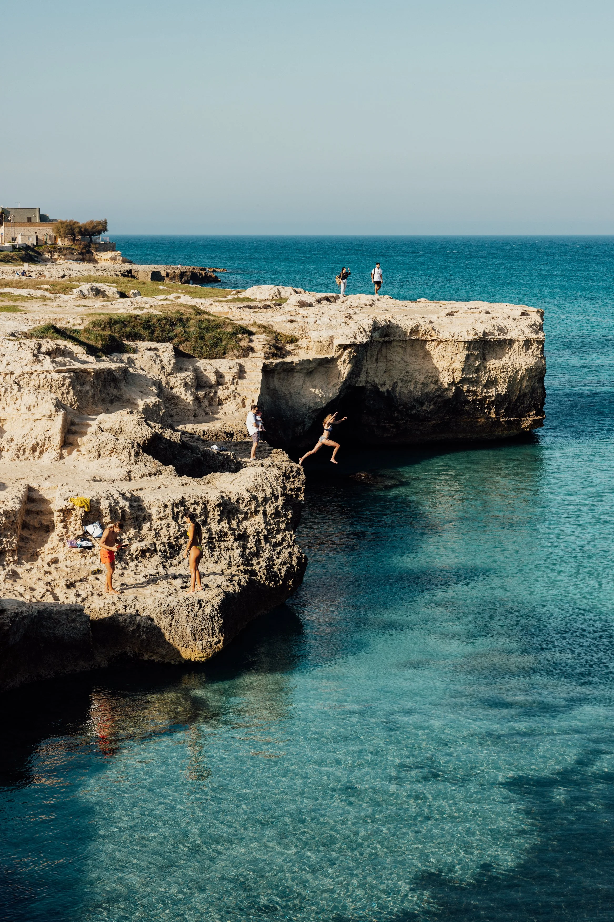 People enjoy a sunny day exploring rocky cliffs and clear blue waters by the coast, with some standing on the cliffs and others jumping into the water.