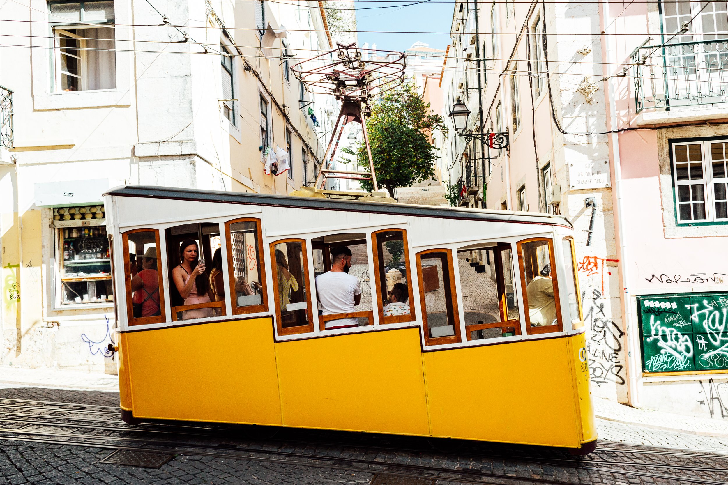 Yellow tram traveling through narrow street with old buildings, some graffiti, and a few passengers inside, one woman checking her phone.