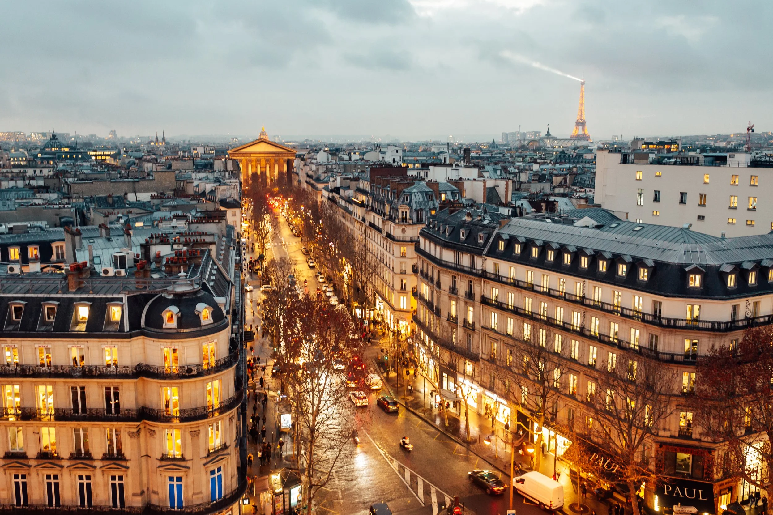 Cityscape of Paris, France at dusk, showing illuminated streets, Haussmann-style buildings, the Arc de Triomphe, and the Eiffel Tower in the background with a cloudy sky.