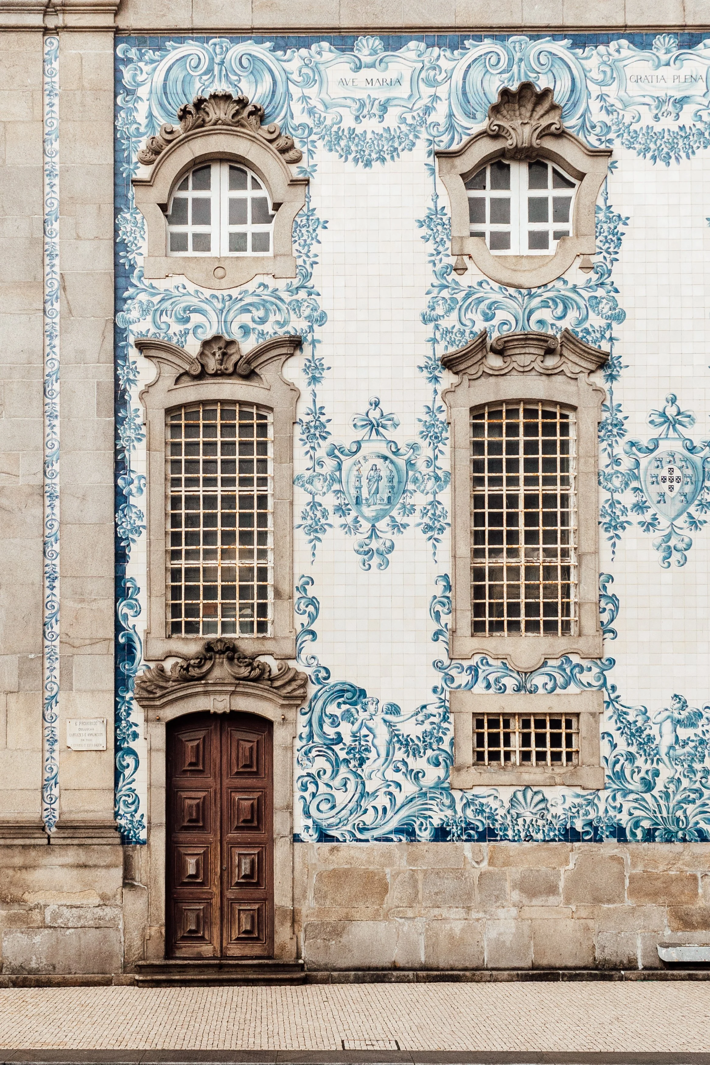 Facade of a building decorated with traditional blue and white azulejo tile artwork, featuring windows with ornate stone frames and a wooden door at the bottom.