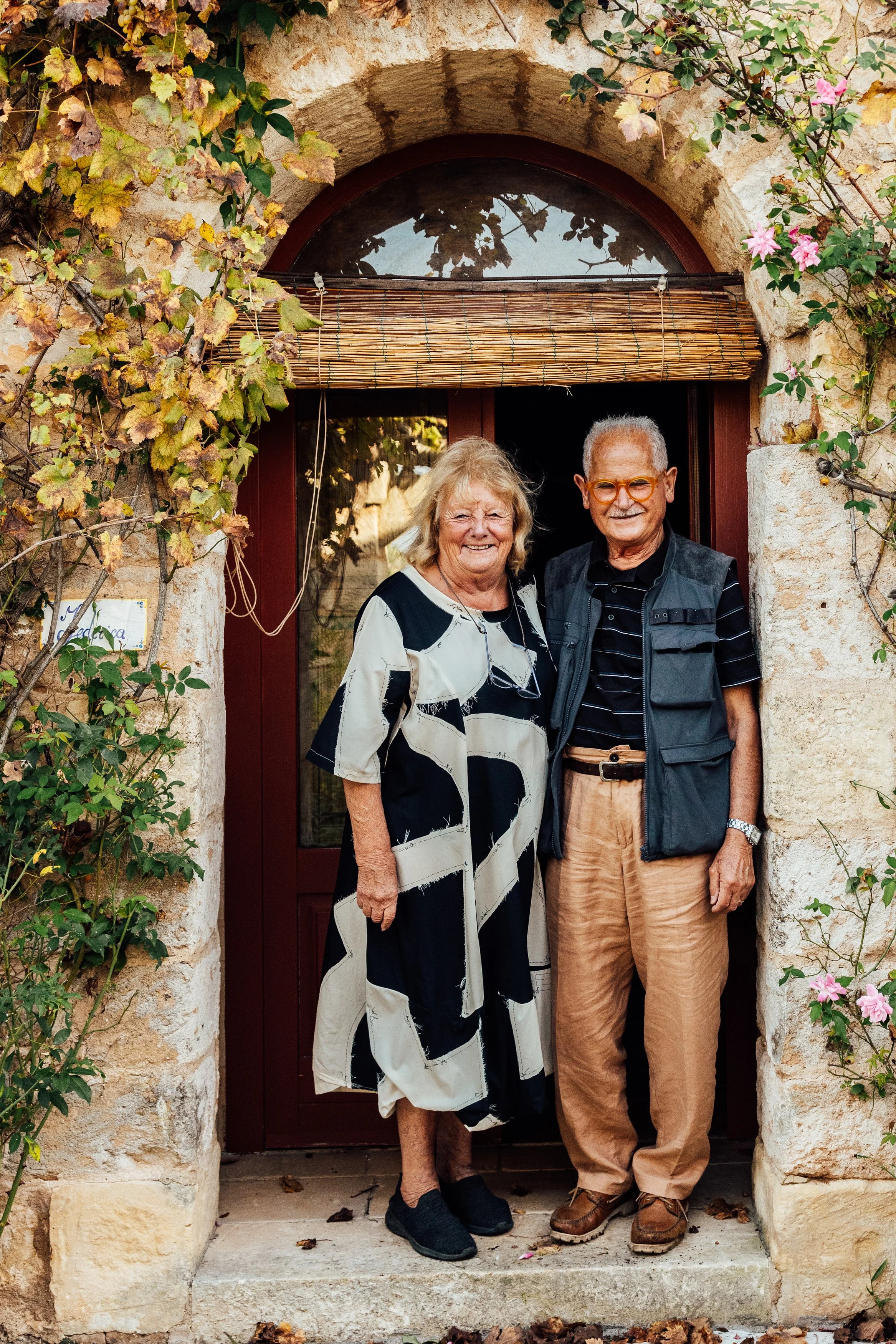 An elderly couple standing together in front of a rustic stone doorway framed with climbing plants and pink roses, smiling at the camera.