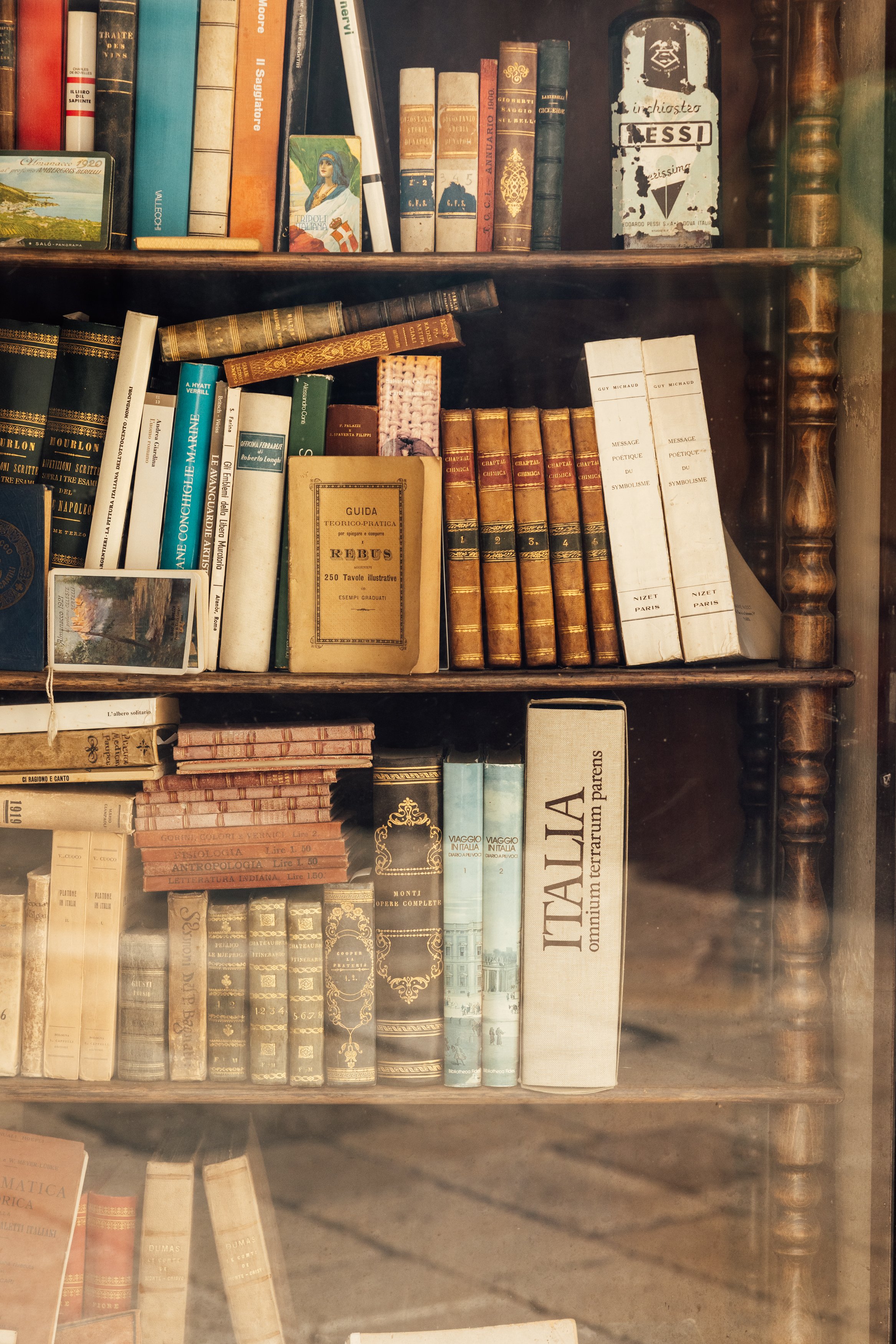 A wooden bookshelf filled with various vintage books, magazines, and small decorative items, including a small postcard.