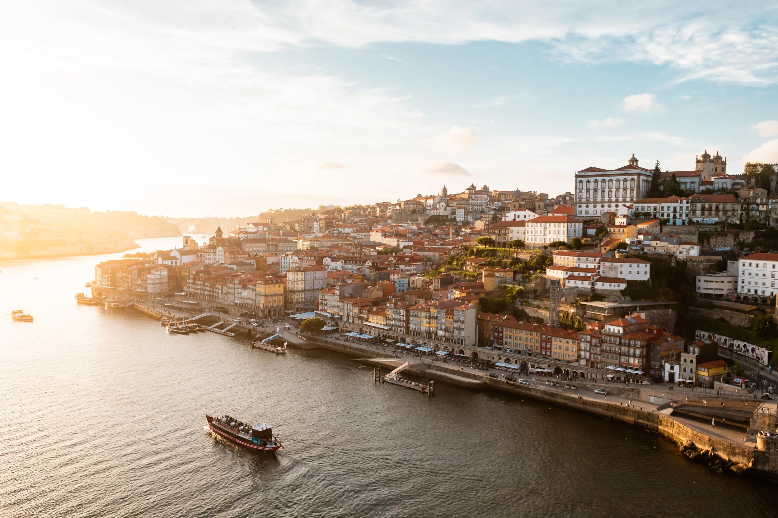 Sunset over a river with boats and colorful buildings on a hillside in a European city.