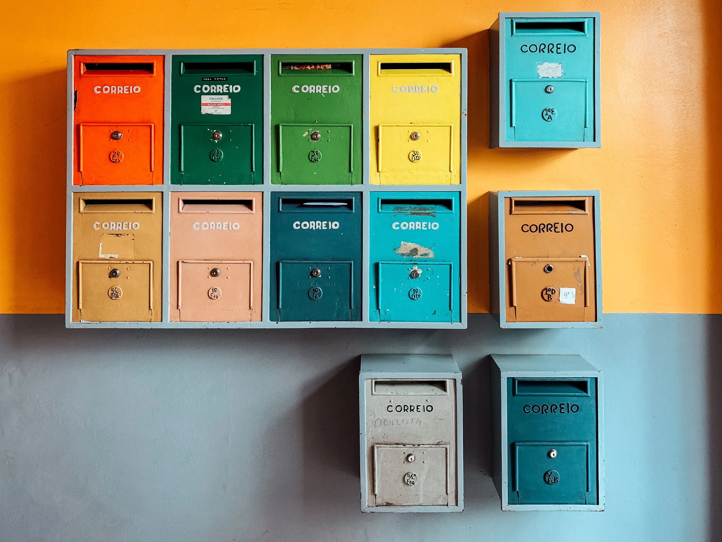 Colorful mailboxes on a yellow and gray wall in a grid pattern. The mailboxes are in orange, green, yellow, beige, pink, blue, brown, white, and turquoise.