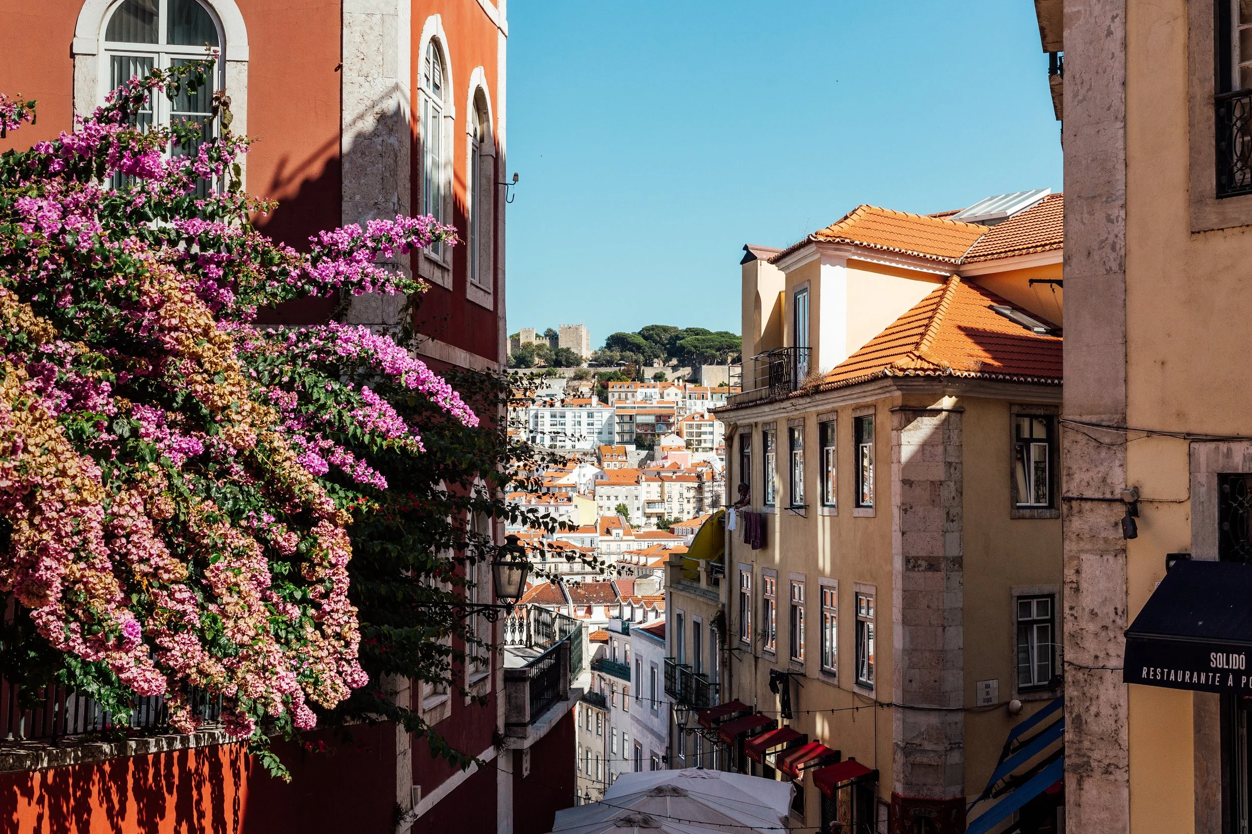 Narrow street in a Mediterranean city with colorful buildings, pink and purple flowering plants on the left, and orange-tiled roofs with a castle on a hill in the background.