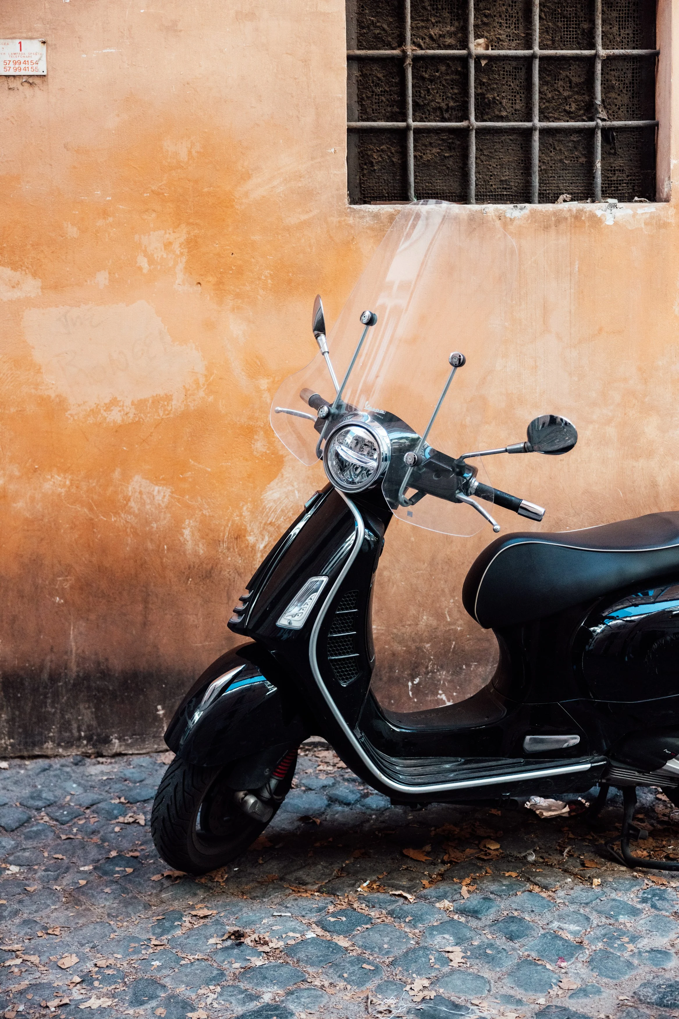 Black scooter with windshield parked on cobblestone street against a weathered orange wall with a barred window.