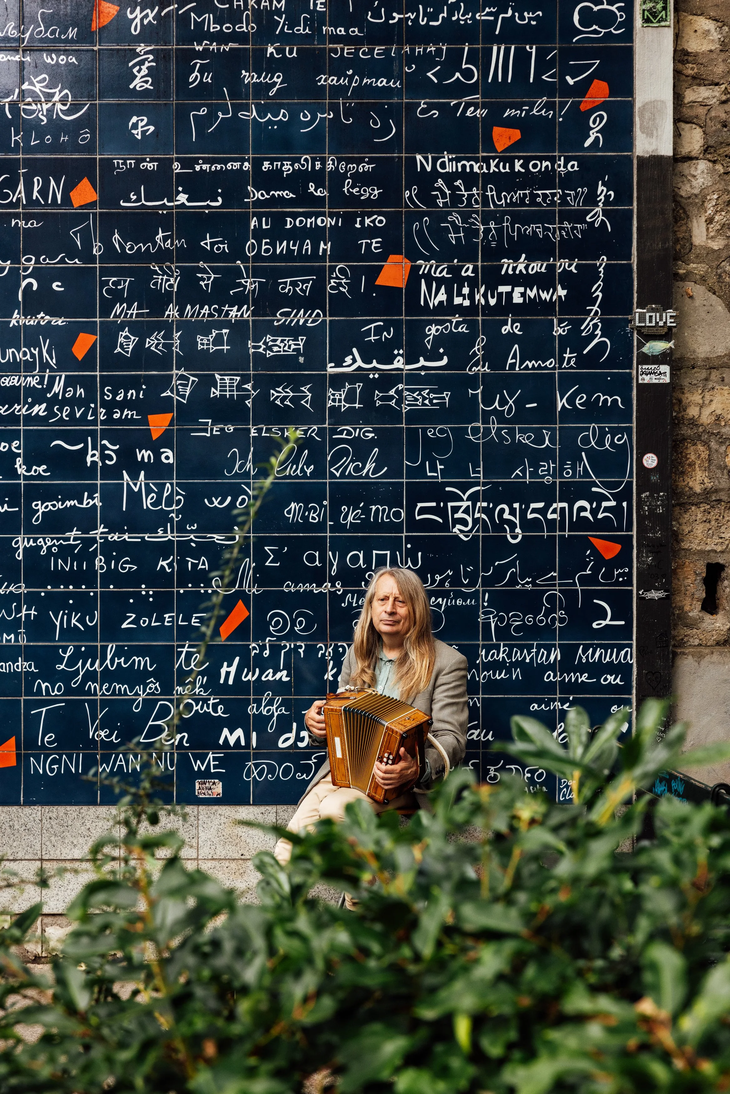 A woman with long blonde hair, wearing a beige blazer, sitting on a stone ledge in front of a large tiled mural with writing in various languages. She is holding an accordion and appears to be out in an urban area surrounded by green bushes.