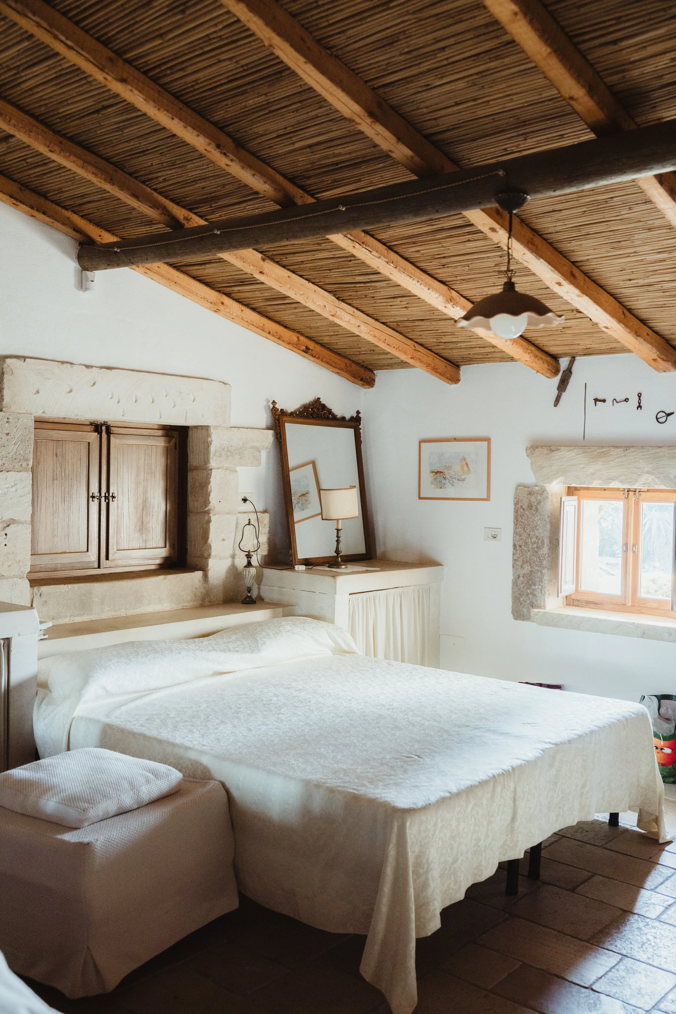 A cozy bedroom with a large bed, wooden ceiling, small window with stone frame, and vintage decor.