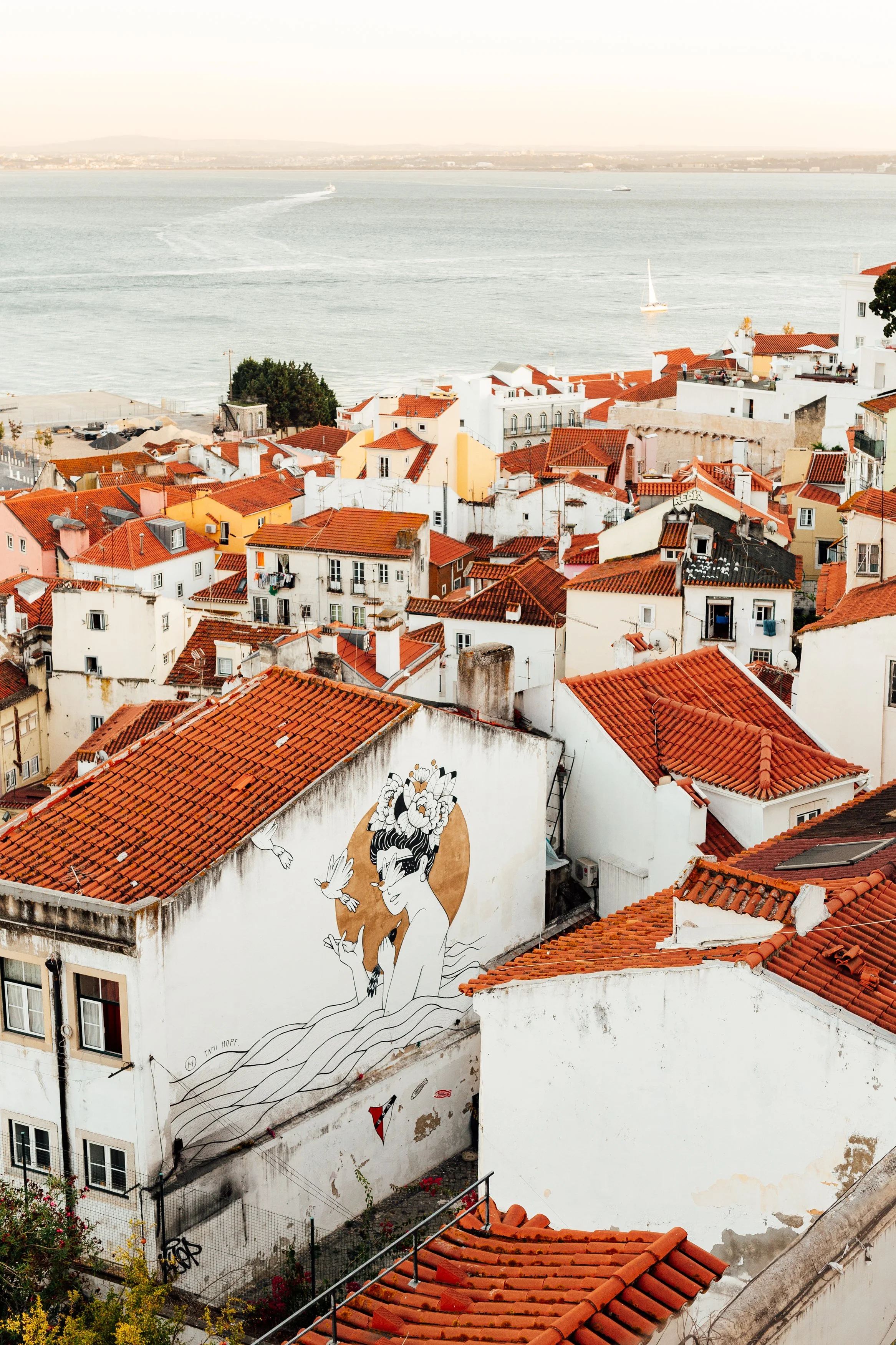 A cityscape of white buildings with red-tiled roofs near a body of water, featuring a large mural of a woman with flowers in her hair and two white birds on a wall.