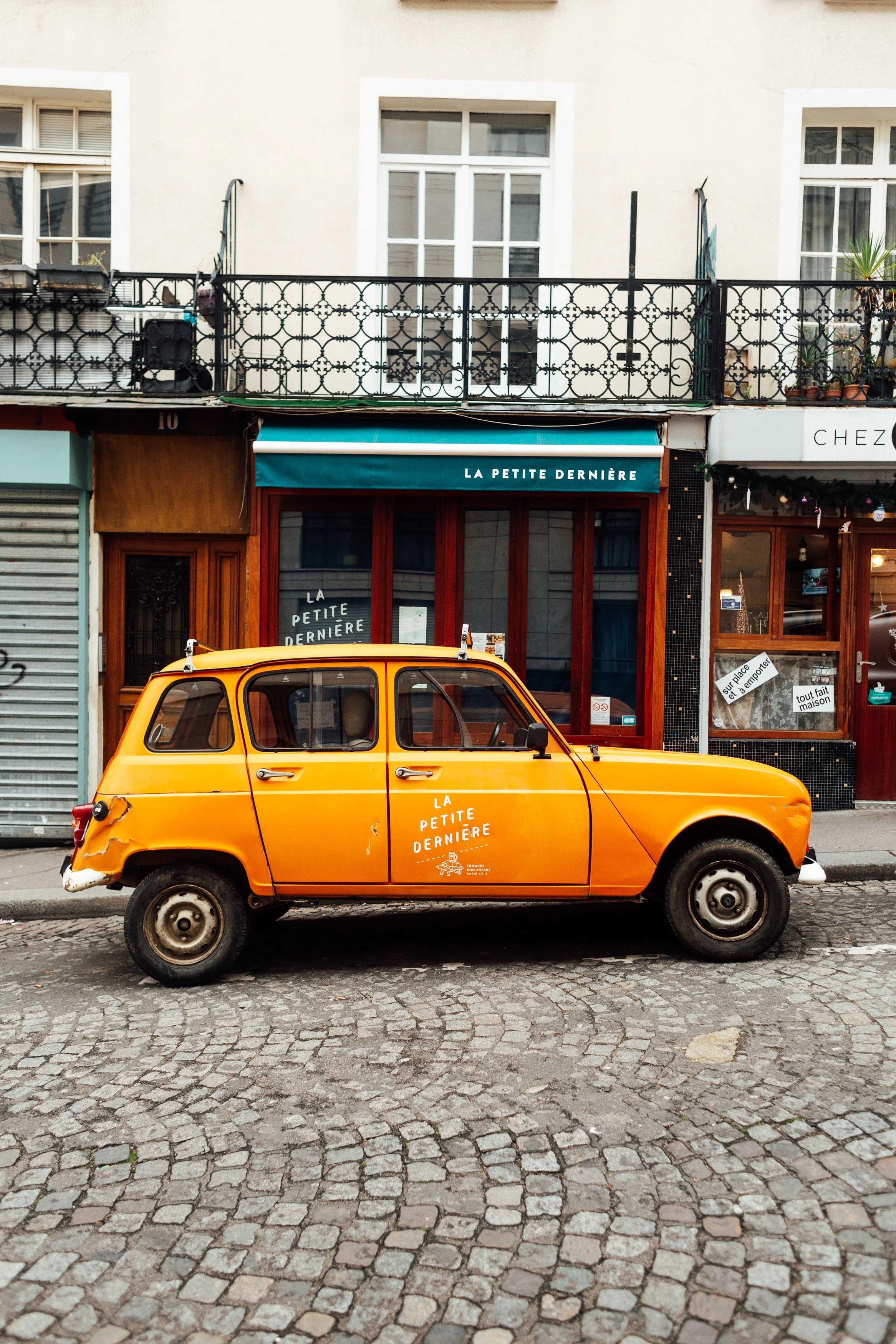 An orange vintage car parked on cobblestone street in front of storefronts with signs and windows, one with an awning reading 'La Petite Dernière'.