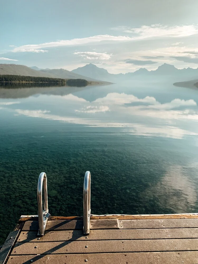 A wooden dock with metal ladders extending into a calm, clear lake that reflects the sky and distant mountains.