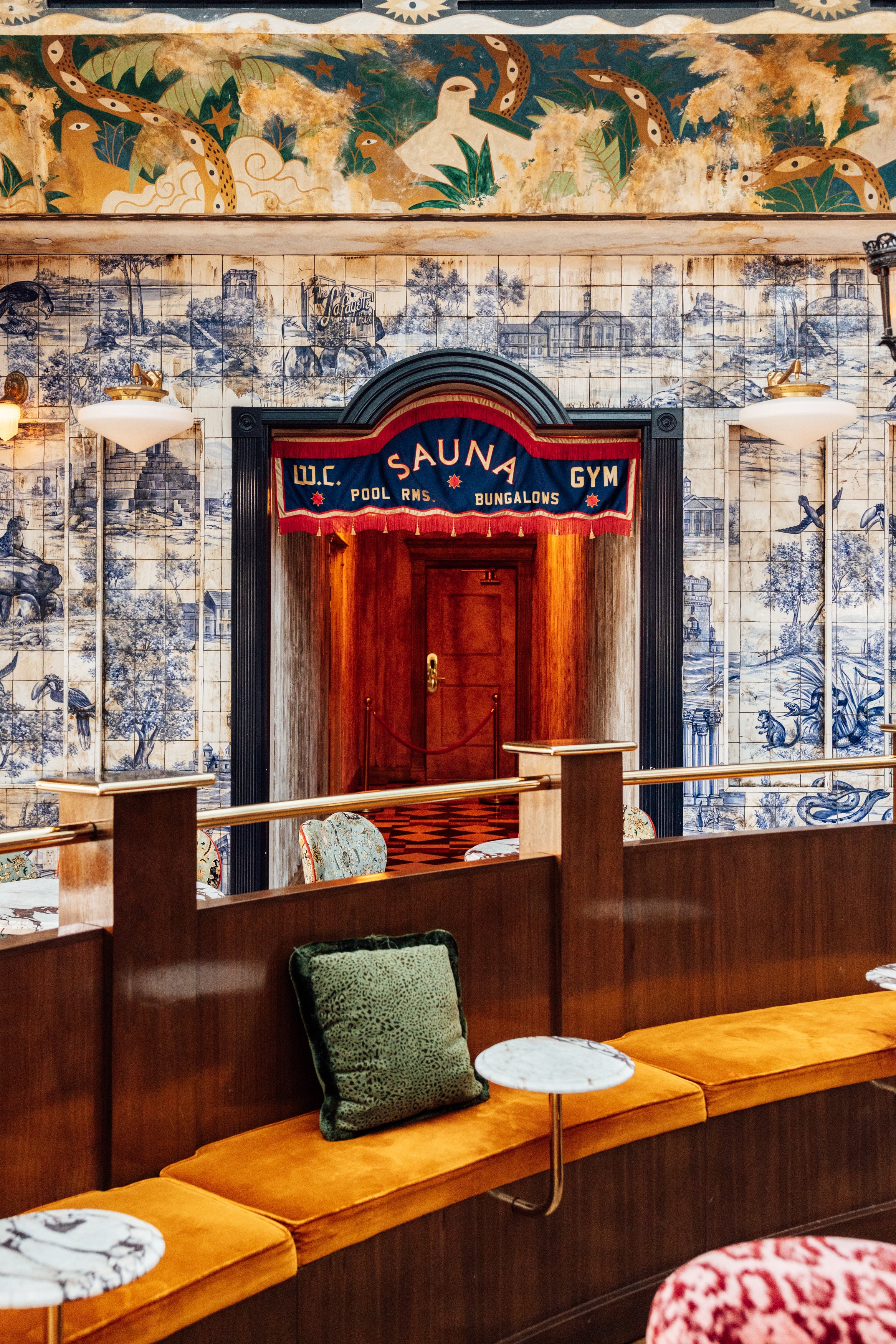 Interior of a vintage lobby with blue and white tile murals, wooden seating with velvet cushions, and a sign for a sauna and gym.