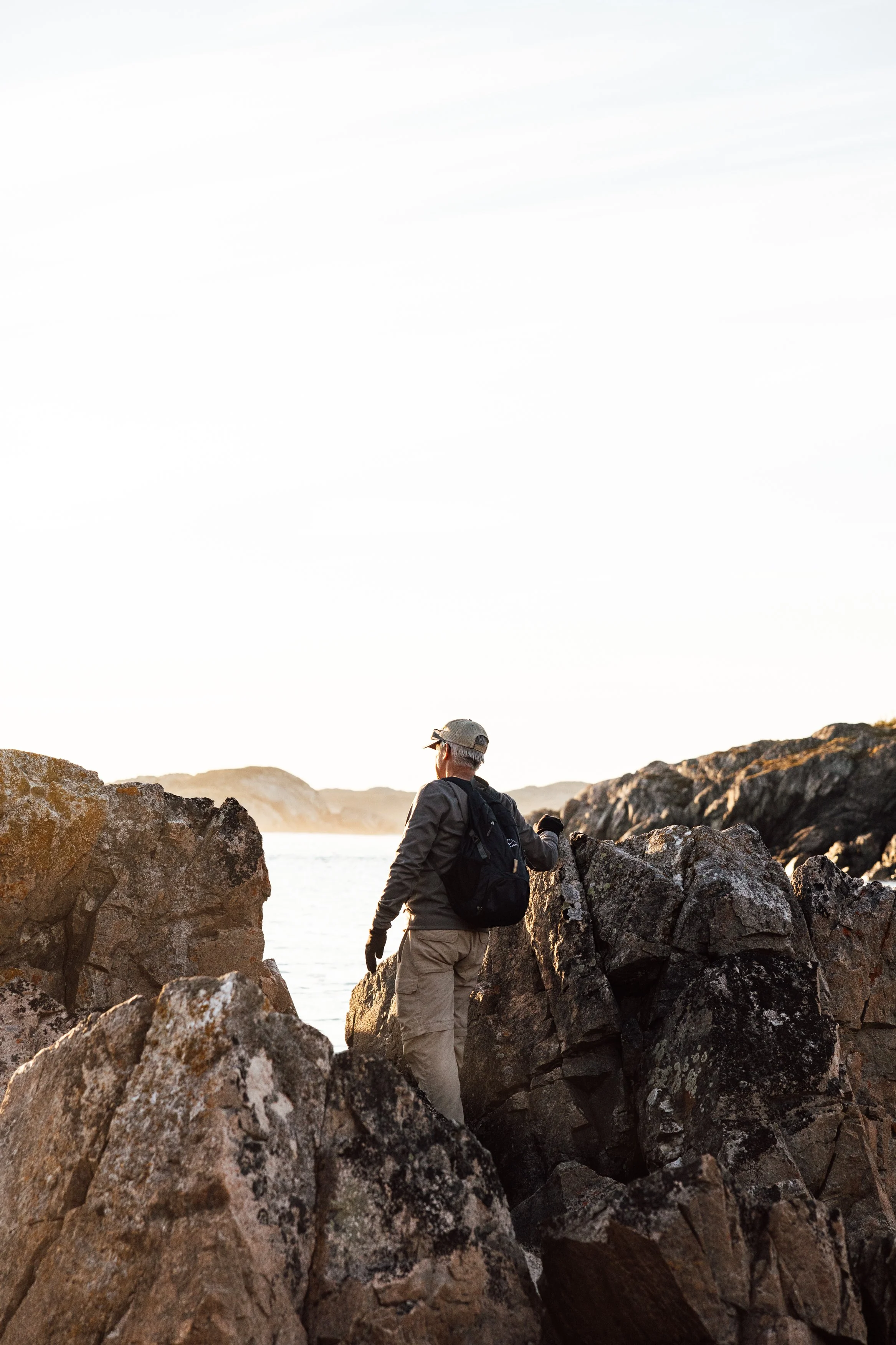 A person wearing outdoor clothing, a cap, and a backpack standing on rocky terrain near water during sunset.