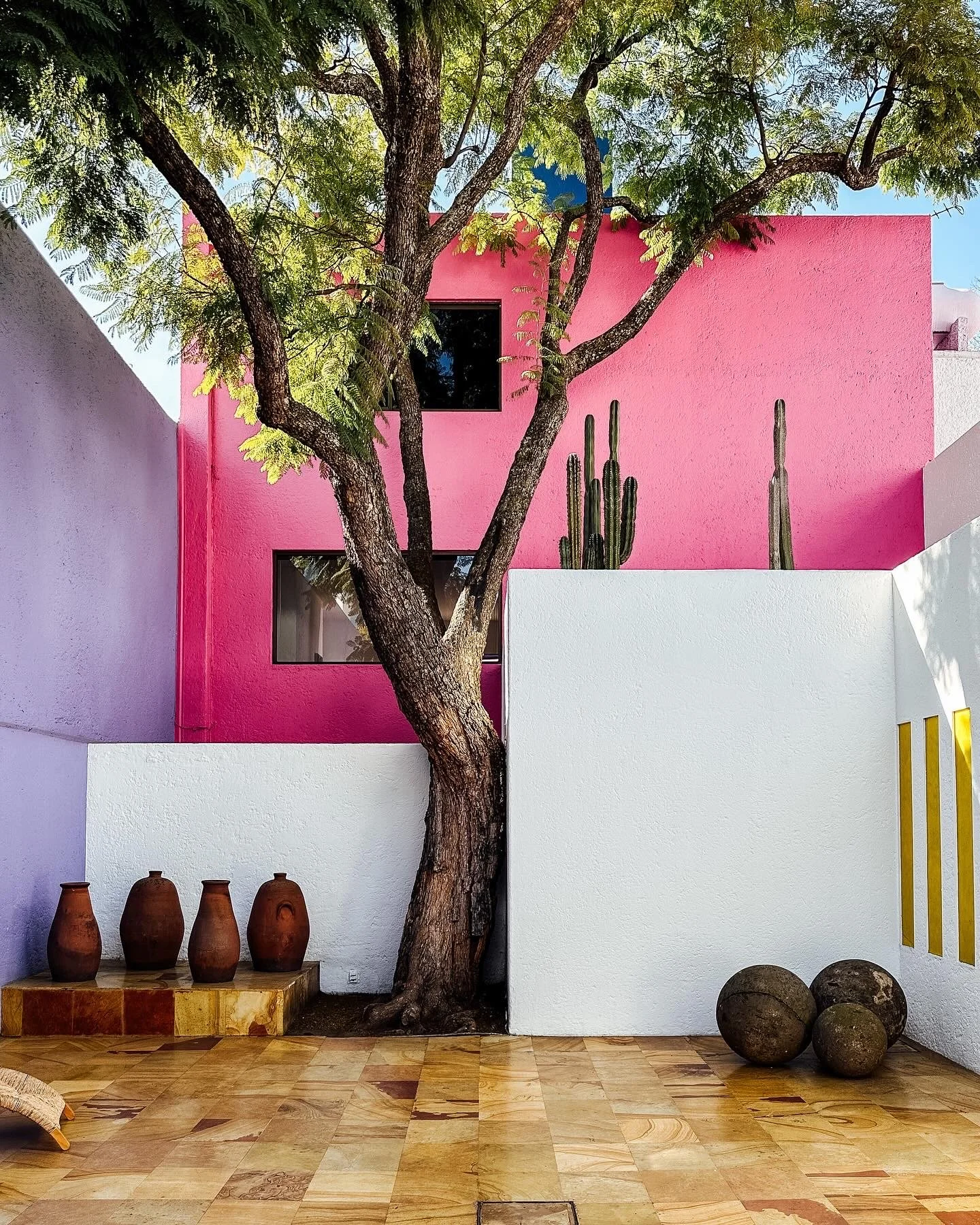 Colorful modern courtyard with a large tree, pink and white walls, cacti, and three large round stones.