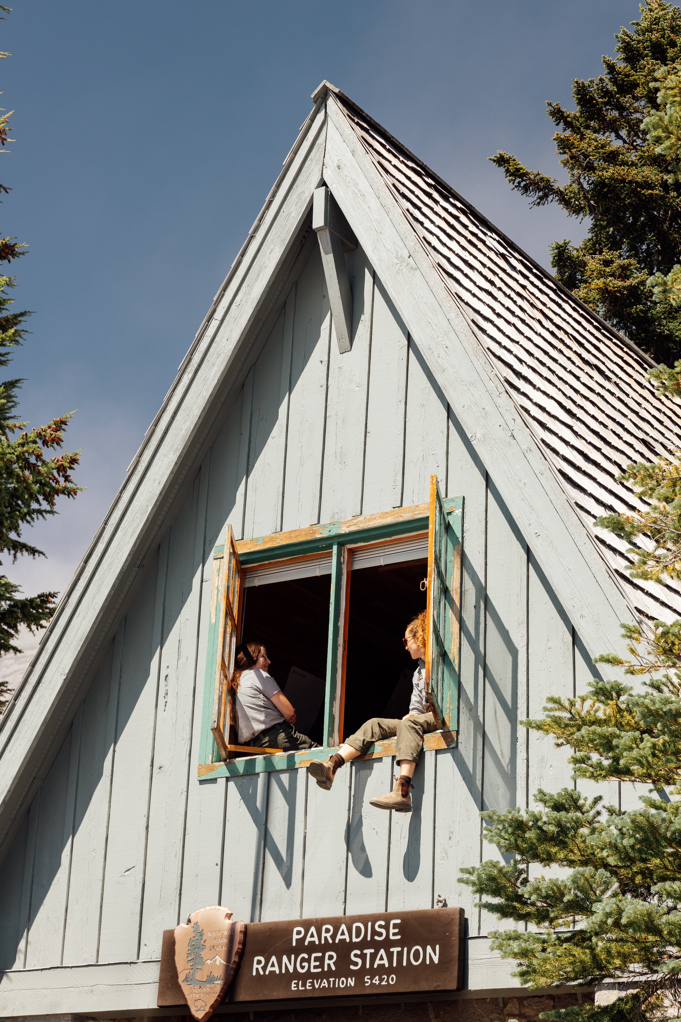 People sitting in an open window of a light blue wooden building labeled 'Paradise Ranger Station, Elevation 5420', with tall evergreen trees on the sides and a partly cloudy sky above.