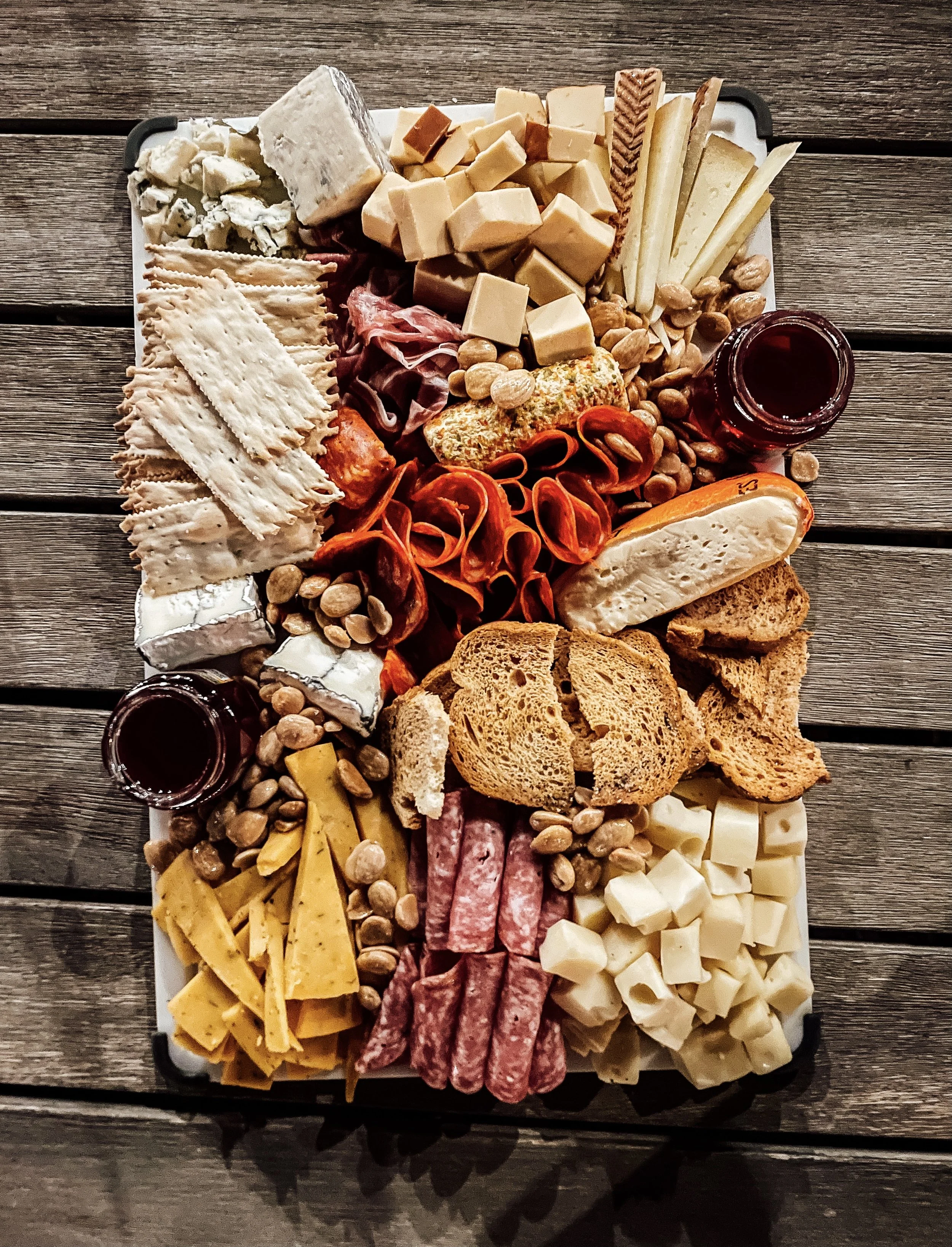A cheese and charcuterie platter with various types of cheeses, cured meats, nuts, bread, crackers, and small jars of jam, arranged on a wooden table.
