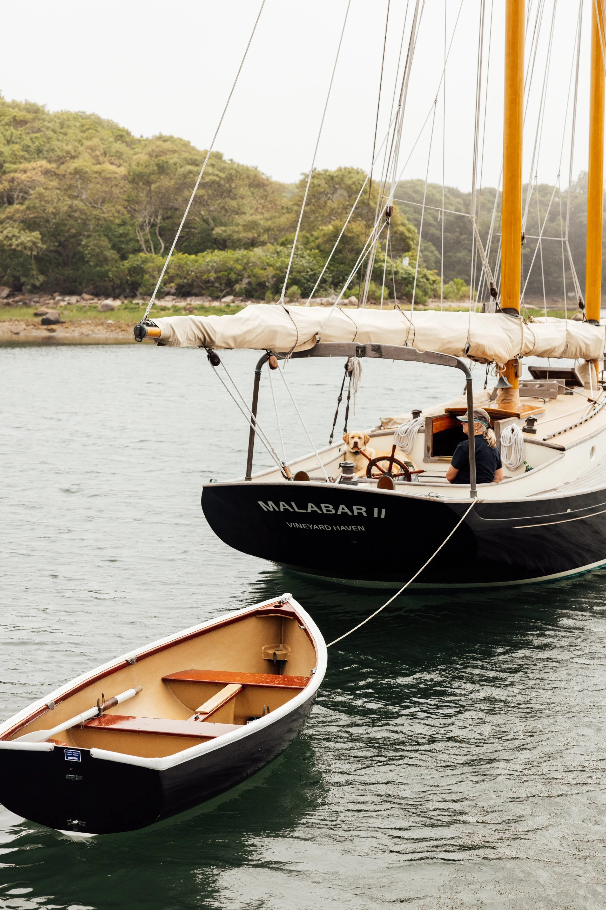 A sailboat with the name Malabar II docked on a calm body of water, with a small rowboat floating nearby. A woman is sitting in the sailboat, steering, accompanied by a dog. In the background are trees and a rocky shoreline.