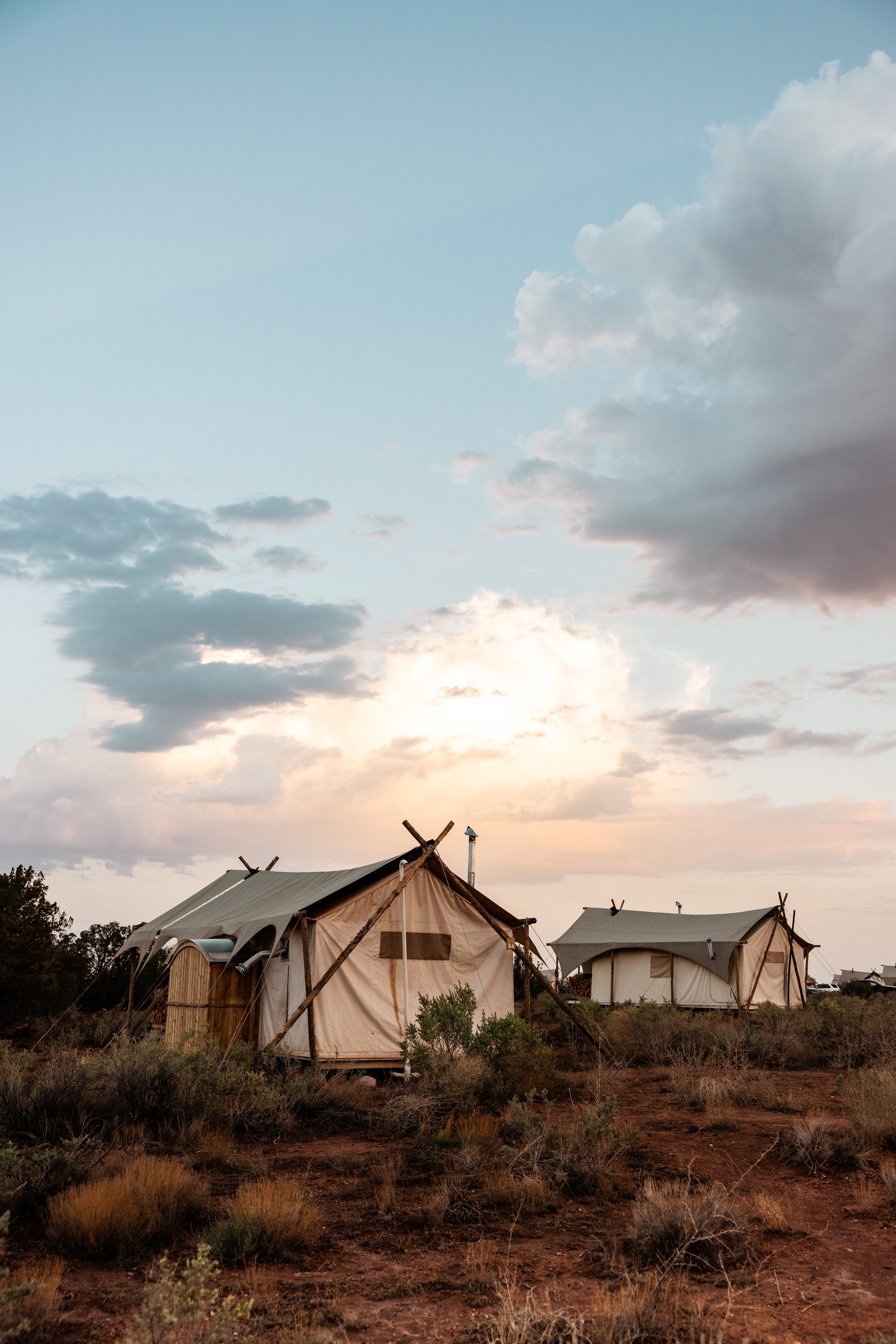 Two white canvas tents set up over a dry, grassy landscape during sunset with clouds in the sky.