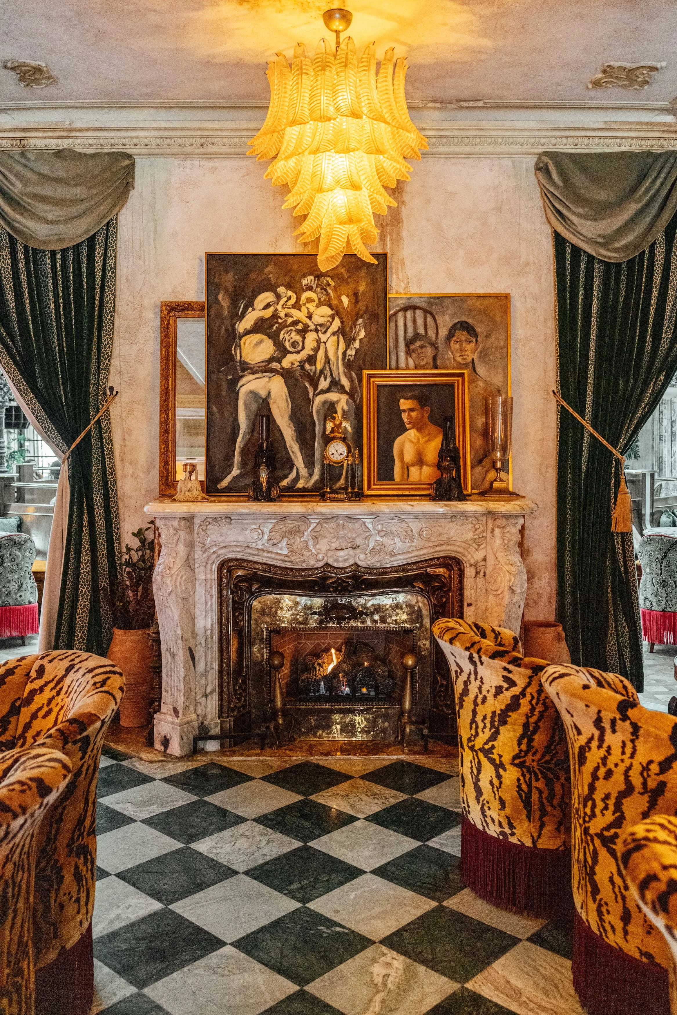 Living room with a marble fireplace, framed artwork, and a large yellow chandelier. Velvet curtains frame windows, and patterned chairs are arranged around a checkered marble floor.