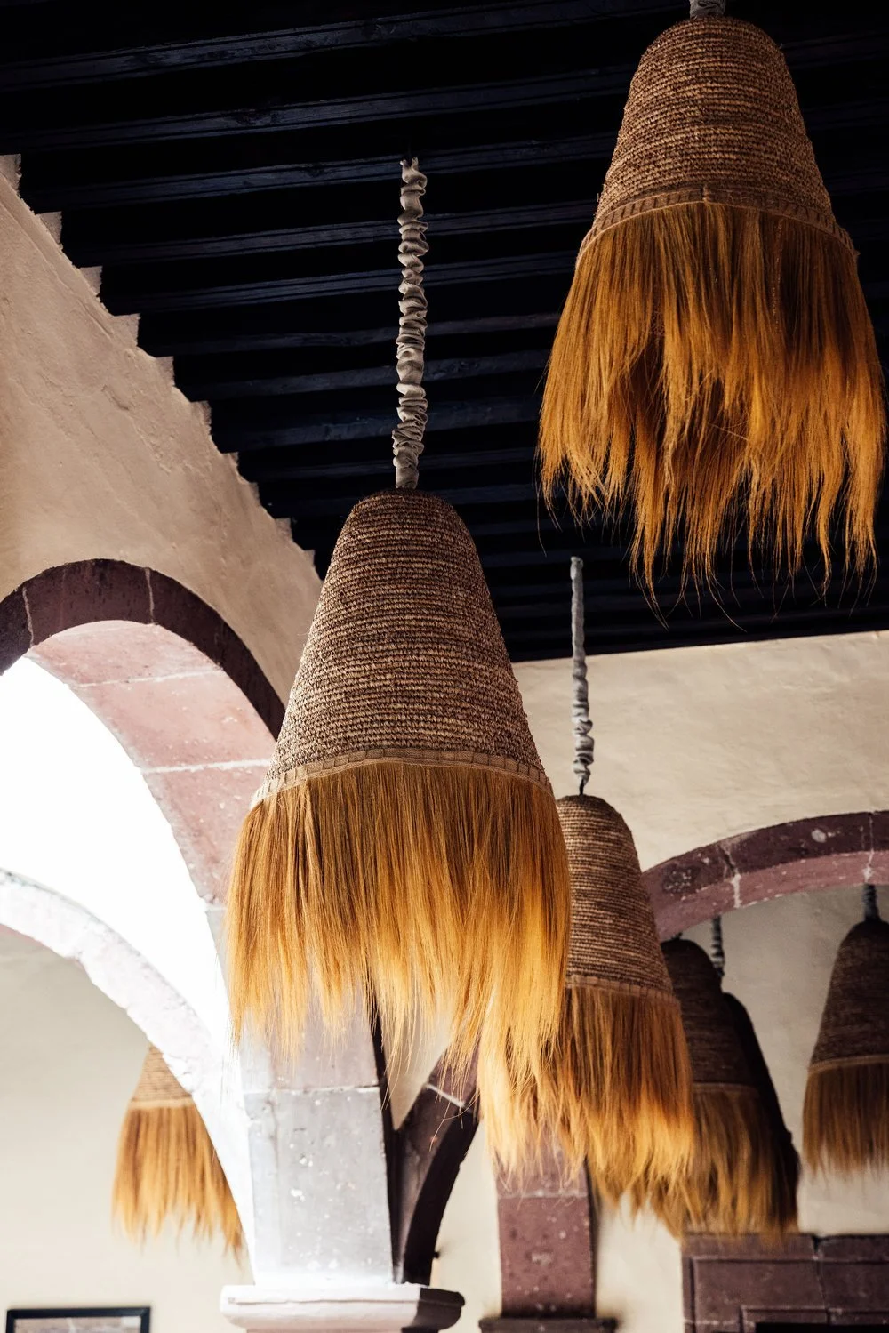 Multiple woven pendant lamps with frayed orange edges hanging from a dark ceiling in a room with arches and white walls.