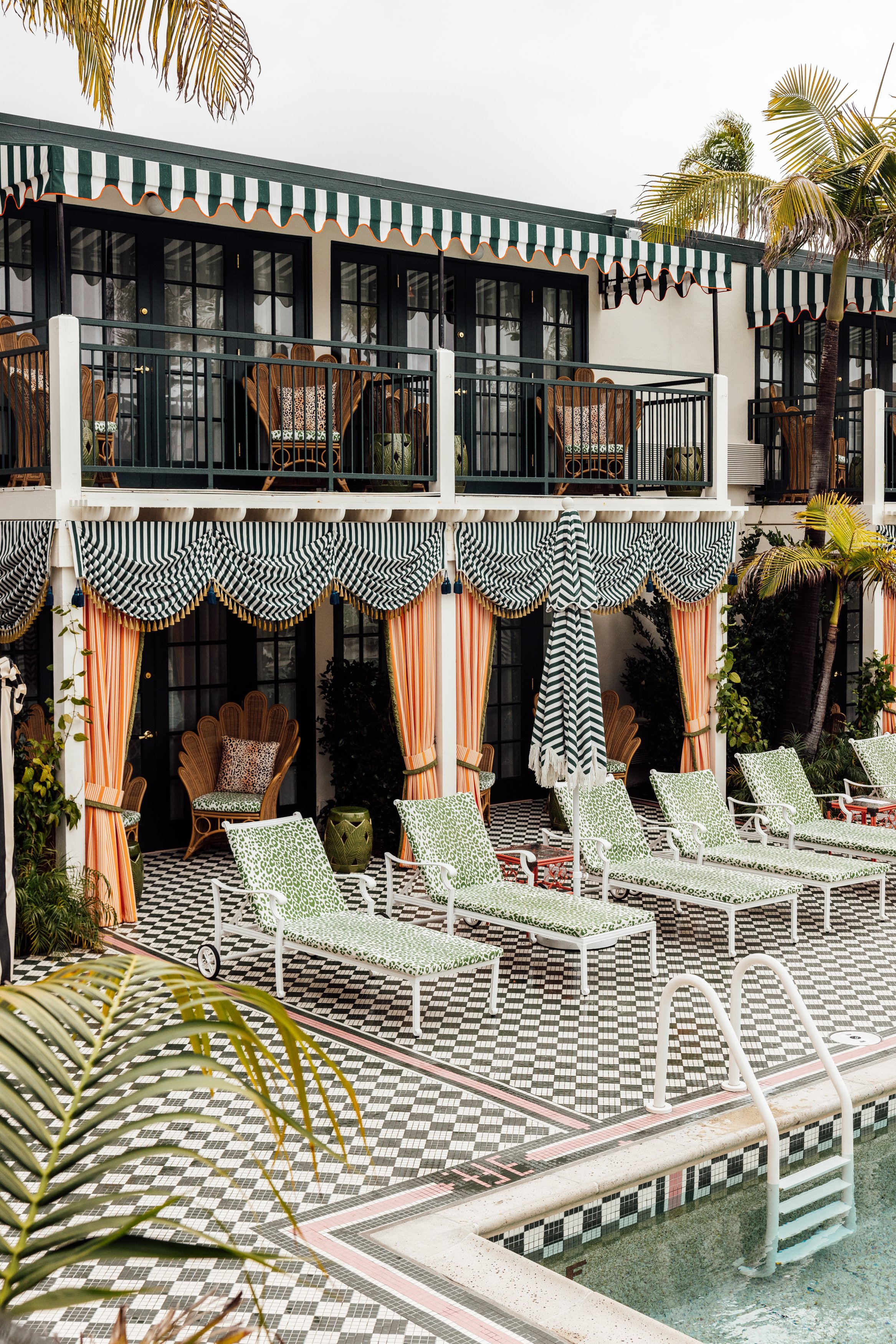 A multi-story outdoor pool area with sun loungers, striped umbrellas, and a balcony with chairs and tables, surrounded by palm trees.