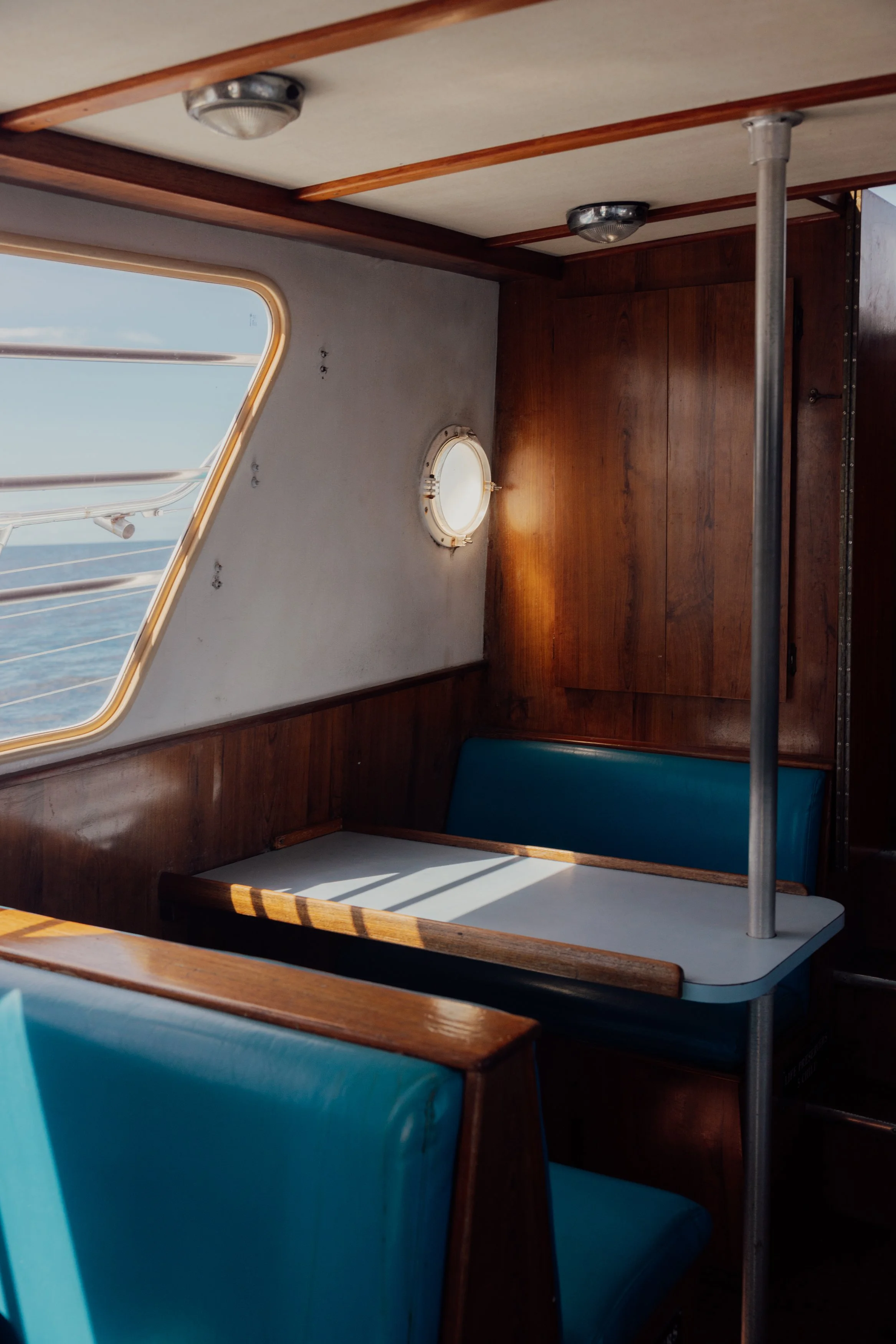 Interior of a wooden boat with a small dining area, blue cushioned seats, a table, a porthole, and a window showing the ocean outside.
