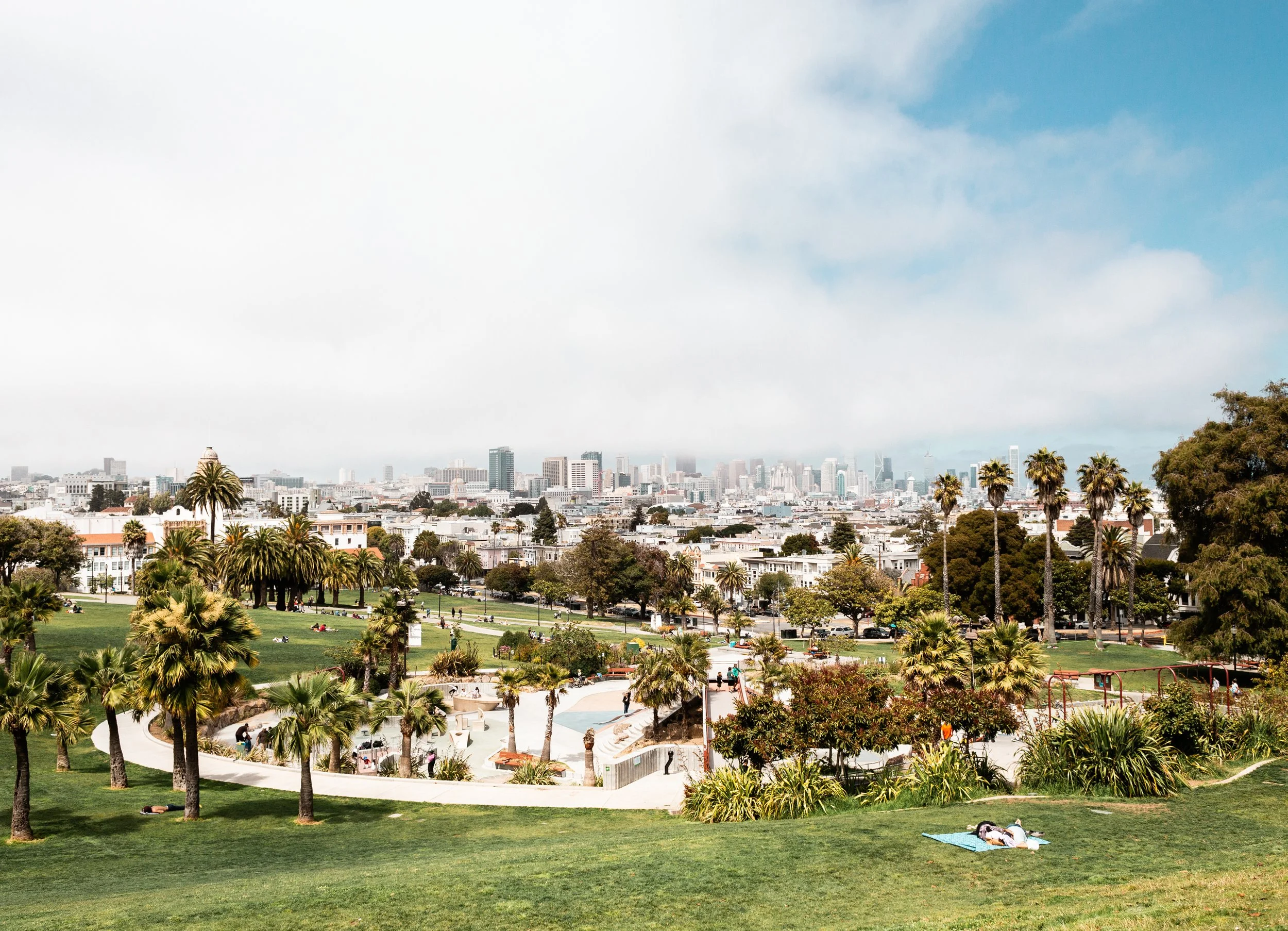 A city park with many palm trees, some people relaxing on grass and walking along paved paths, with a skyline of tall buildings in the distance under a partly cloudy sky.