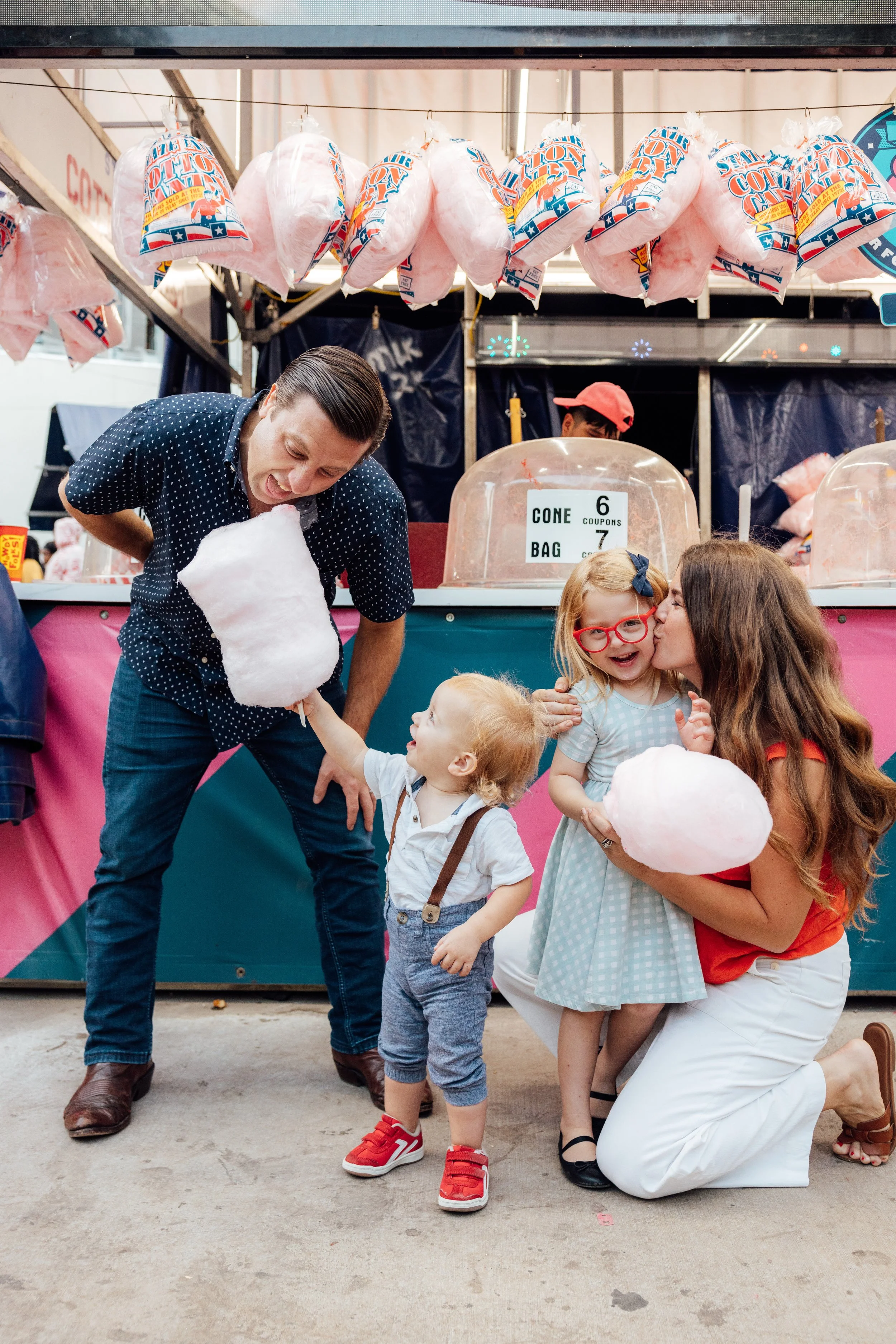A family at a cotton candy stand, with two young children being given cotton candy by a man, and a woman kissing a young girl while holding cotton candy.
