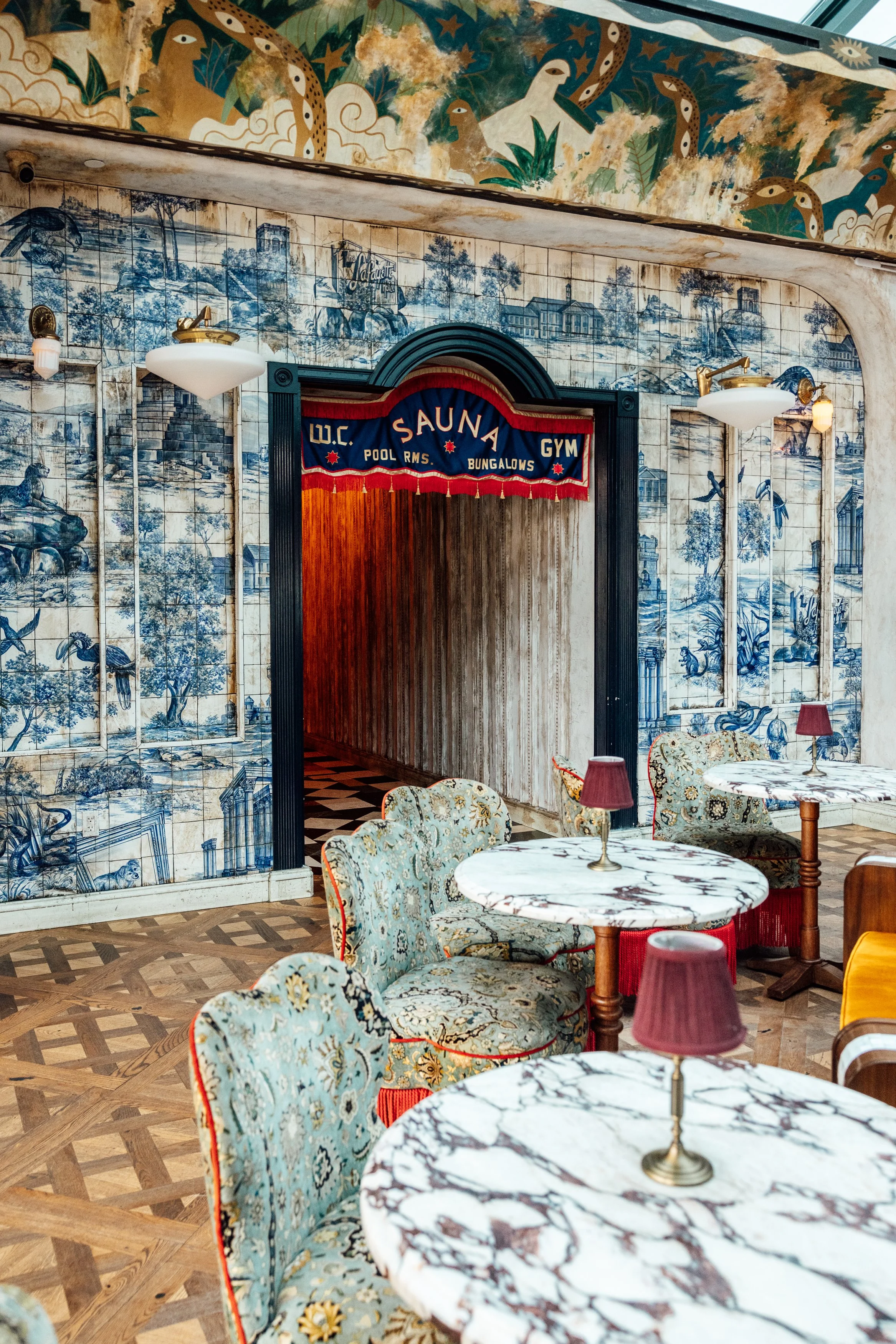 Interior of a vintage-style cafe with marble tables, patterned upholstered chairs, and blue and white wall murals featuring landscapes and birds. A sign over an arched doorway reads 'Sauna' with additional smaller text.