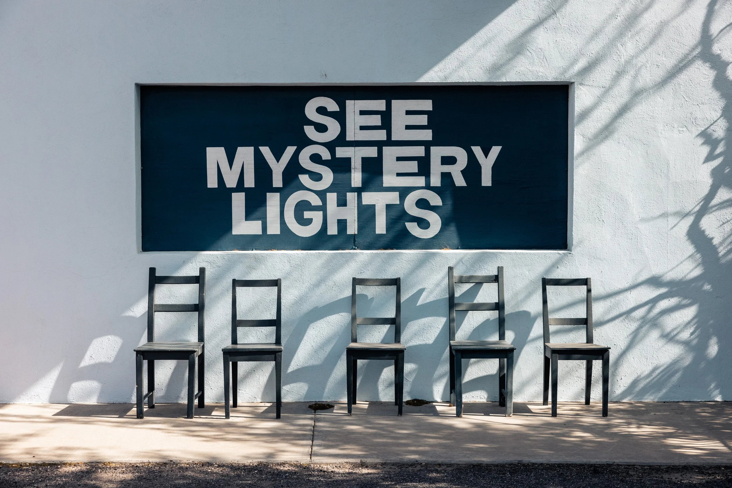 Five black chairs aligned against a white wall beneath a sign that reads 'See Mystery Lights' with shadows cast on the wall.