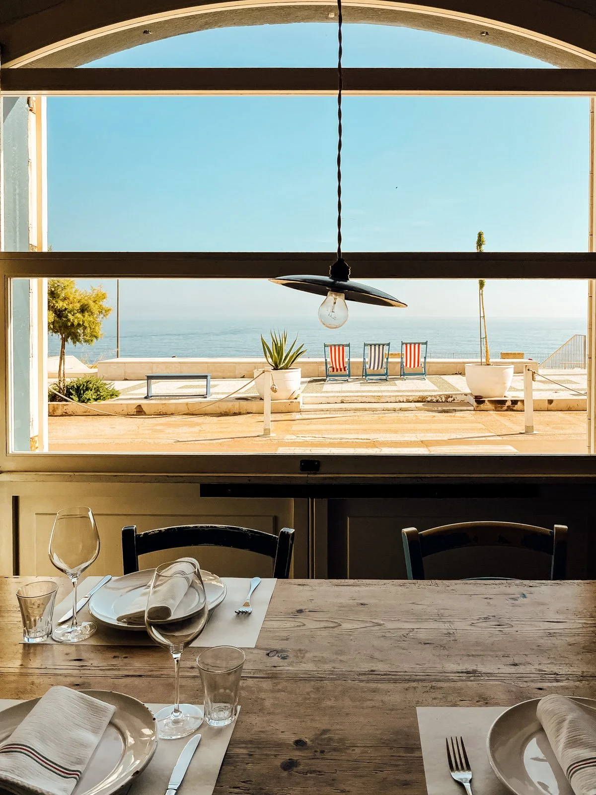 A view from inside a restaurant looking out a window to an outdoor patio with chairs, potted plants, and the ocean in the background.