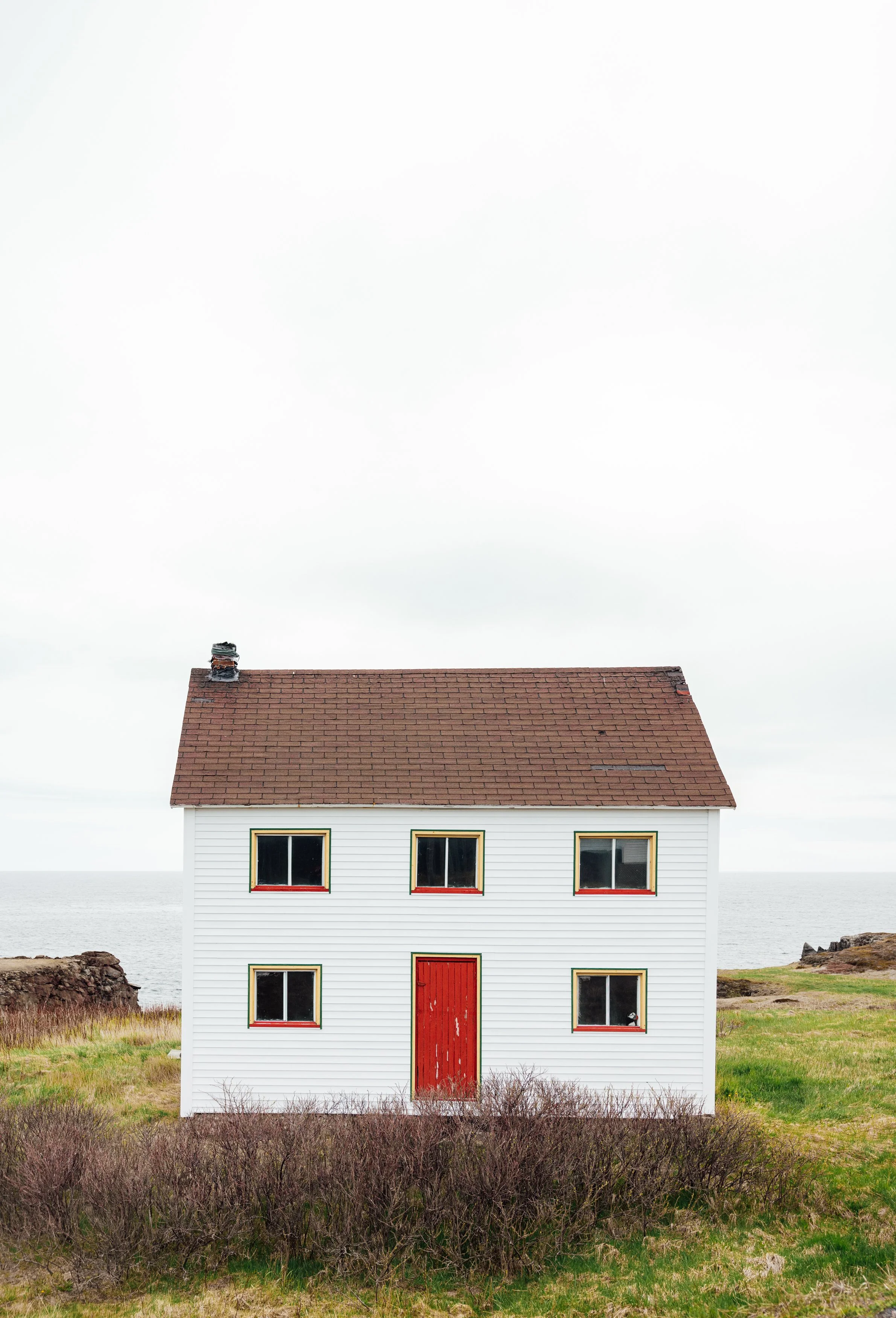 A white two-story house with a red door and red-trimmed windows, situated on a grassy area near the coast under a cloudy sky.