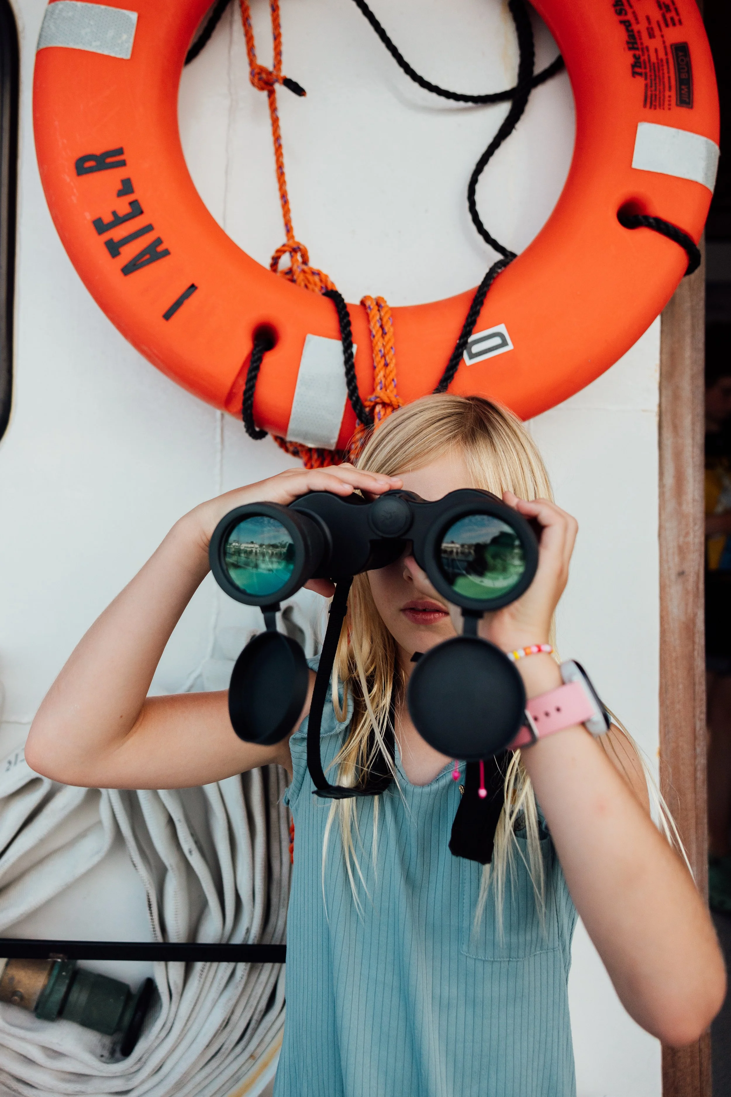 A young girl with blonde hair looking through binoculars, standing in front of a white wall with an orange life preserver mounted above her. The life preserver has black and white clips and orange and black ropes.