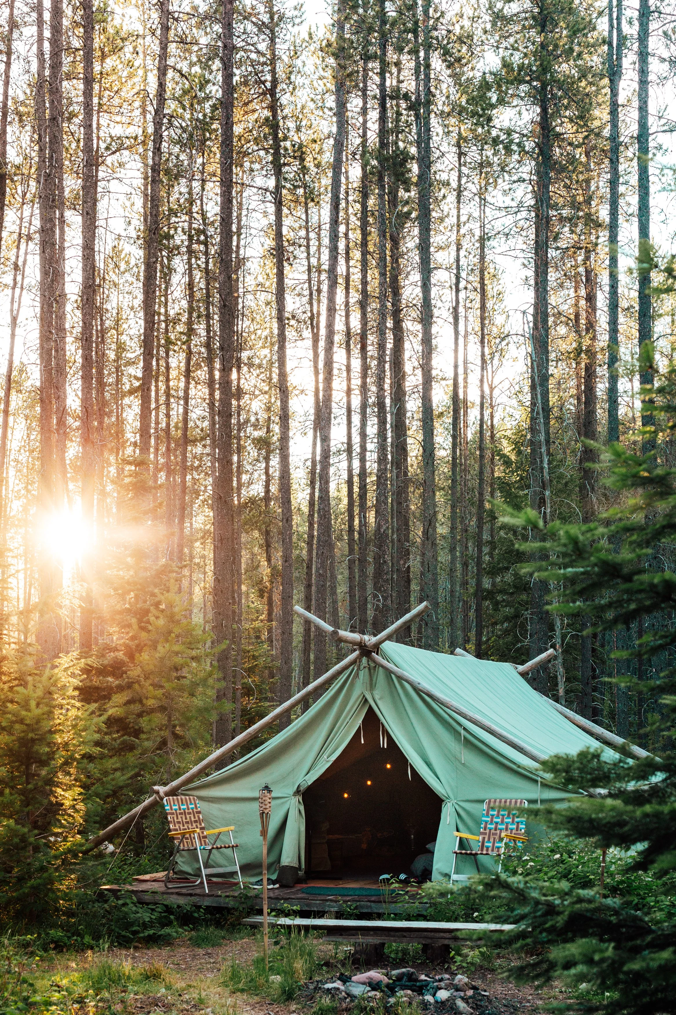 A green camping tent with two chairs outside, set in a forest during sunset.