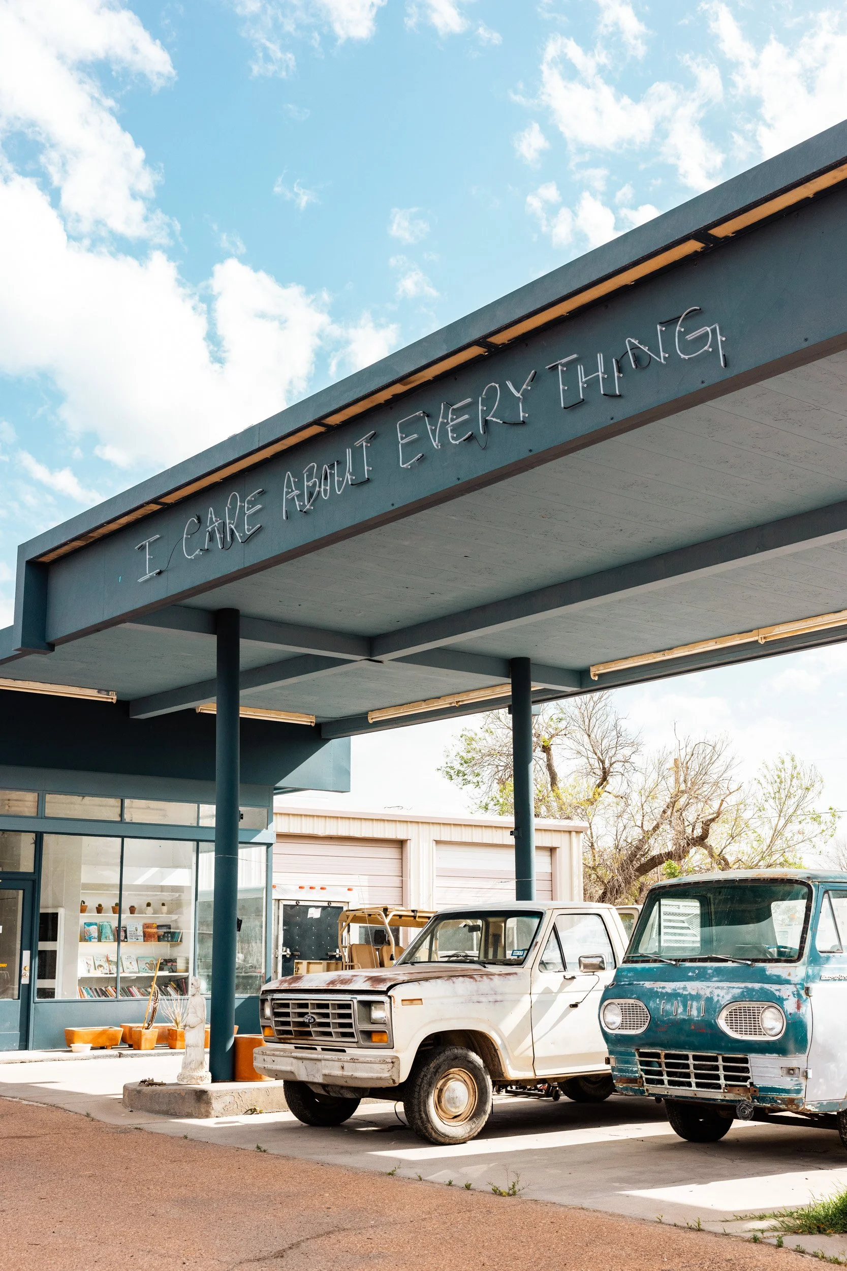 Vintage cars parked in front of a small store with a blue exterior and a sign that reads, 'I CARE ABOUT EVERYTHING,' under a partly cloudy sky.