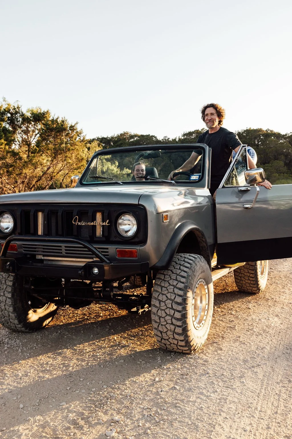 Two people enjoying a ride in a vintage off-road vehicle, with one standing in the passenger seat. The vehicle is parked on a dirt surface with a background of trees and a clear sky.