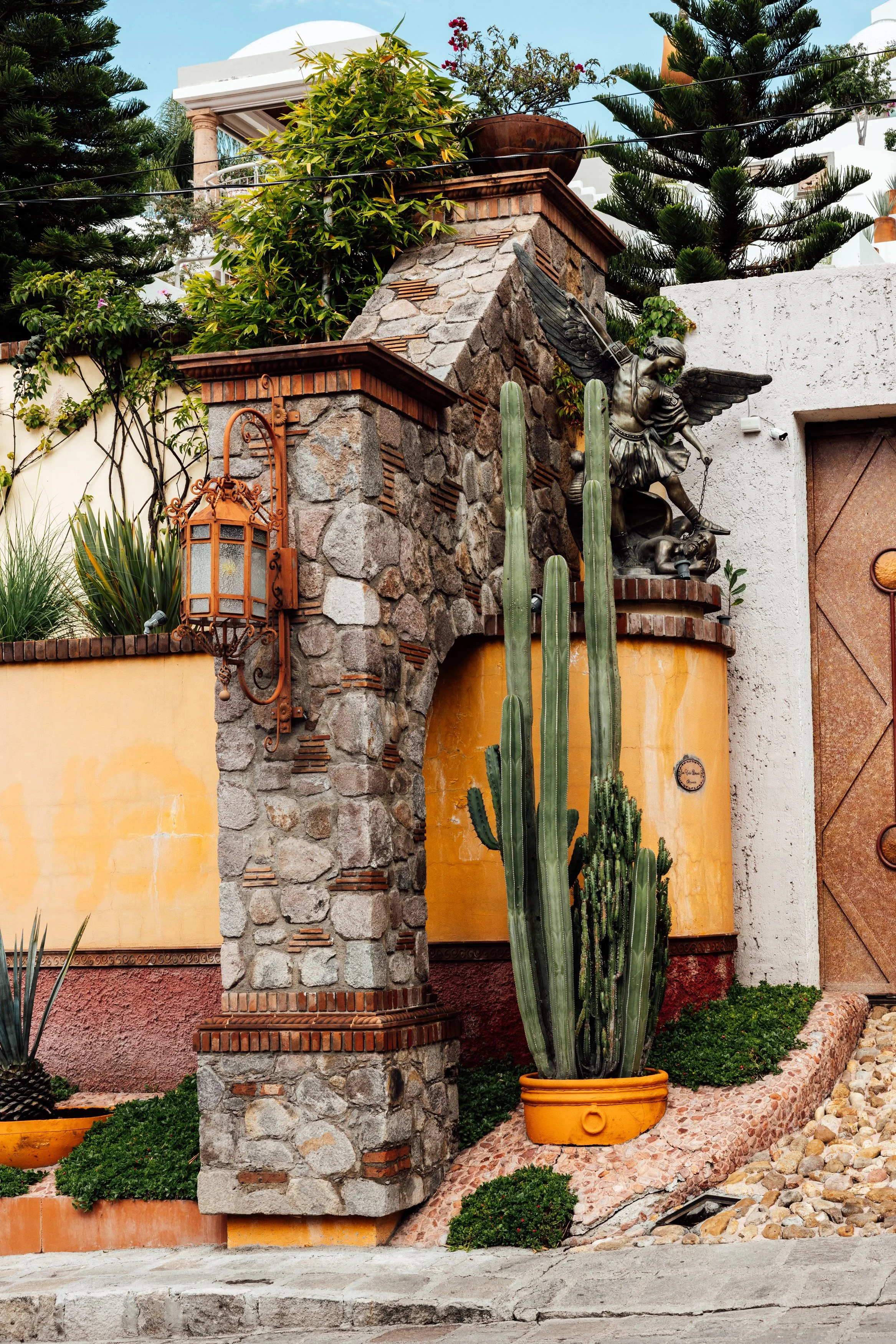 Decorative exterior wall with a stone pillar, a cactus in an orange pot, and a bronze sculpture of an angel with wings, holding a bow and arrow, perched above the wall.