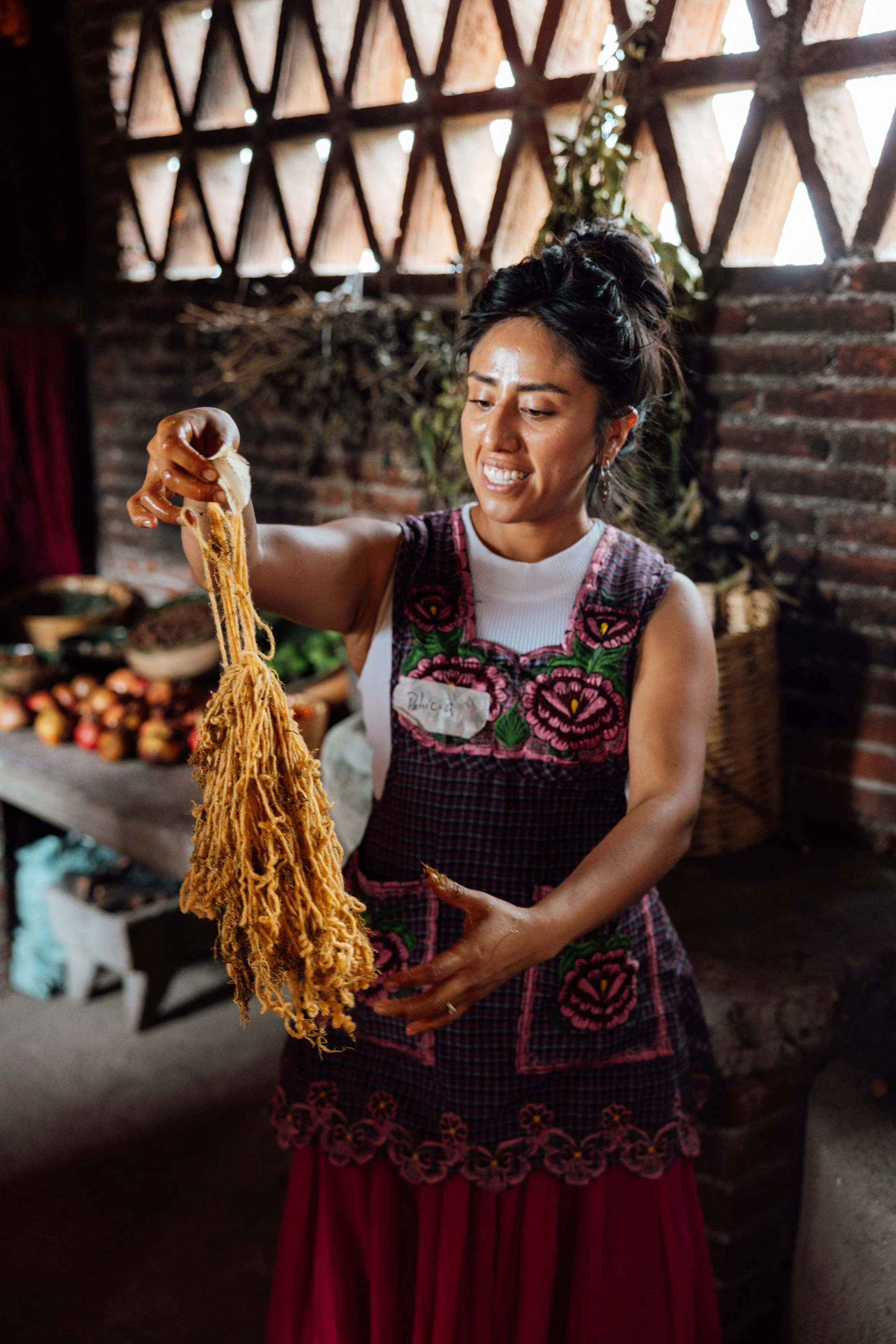 A woman holding a bunch of freshly made, spun yarn in a rustic indoor setting with a brick wall and wooden window in the background.