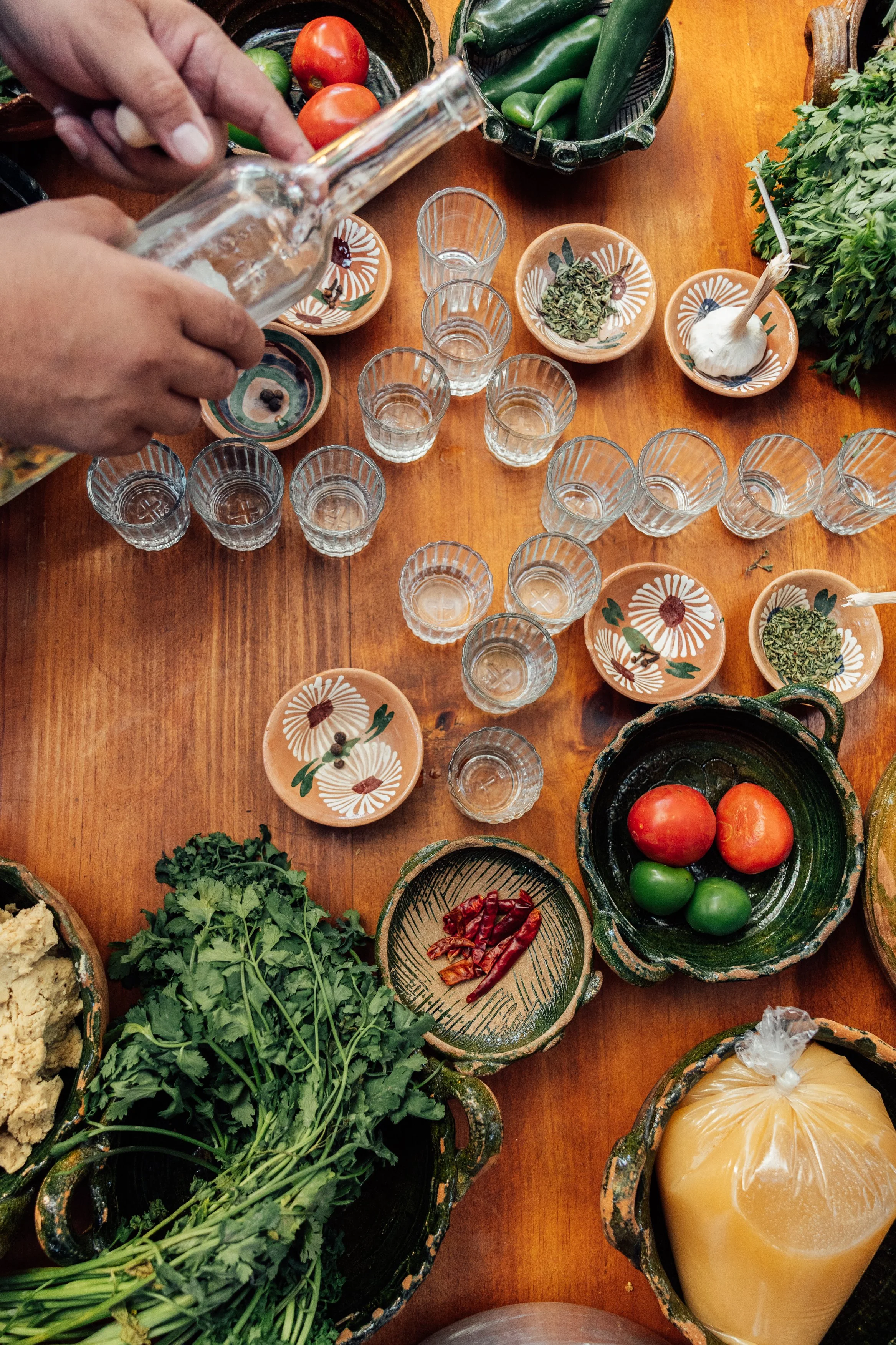 Person pouring clear liquid into small glass shot glasses on a wooden table with various fresh vegetables, herbs, and bowls with dried chili peppers, garlic, and other ingredients.