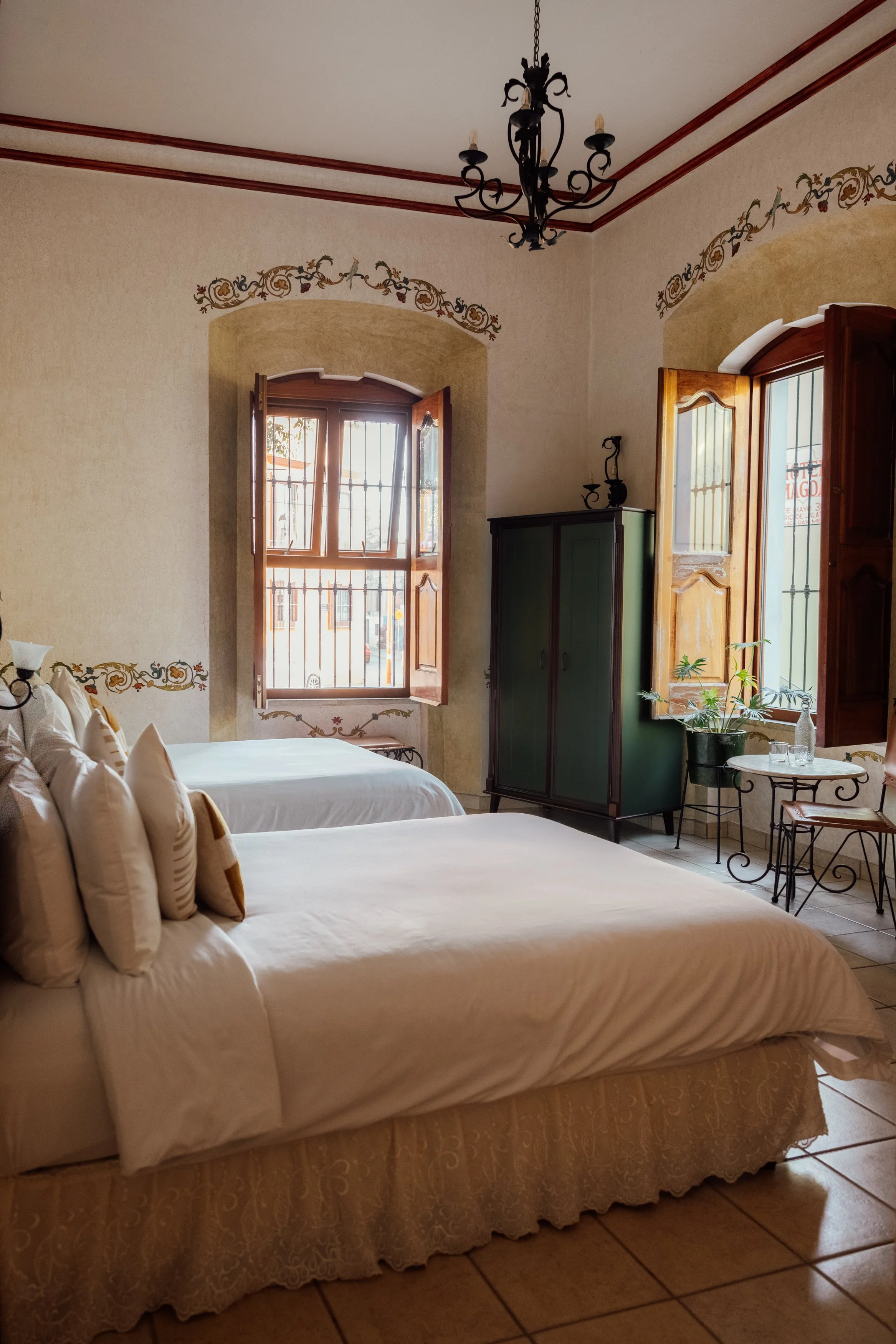 Room with two beds, wooden window shutters open, green wardrobe, bedside table with glass bottles, potted plant, wrought iron furniture, chandelier, decorative wall trim, tiled floor, sunlight streaming in.