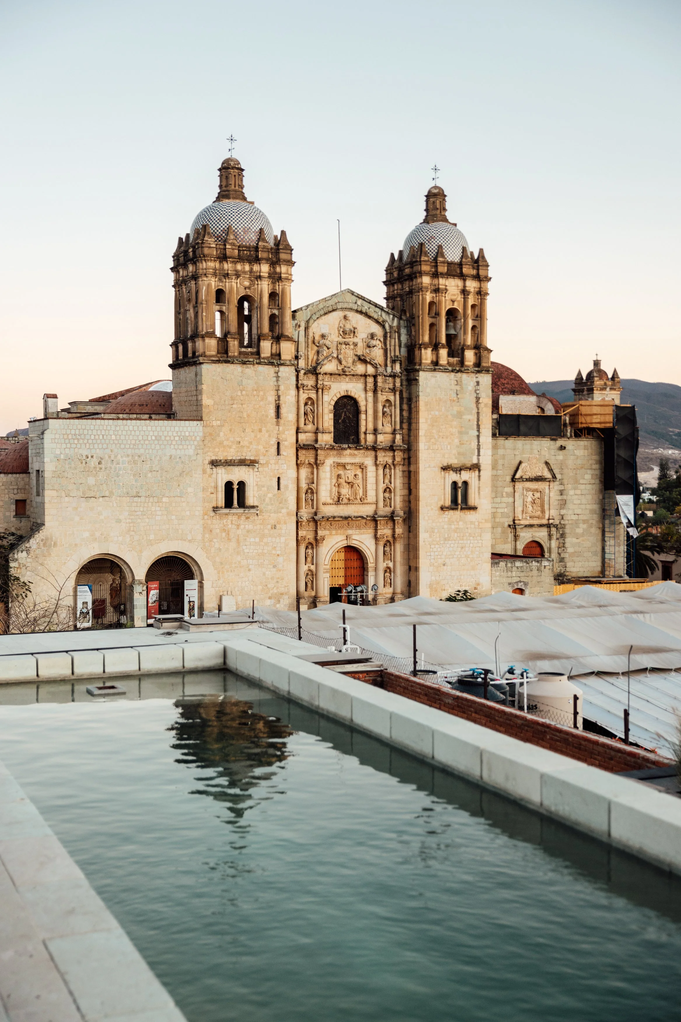 Historic stone church with two bell towers, ornate facade, and sculpture details, seen from above near a rooftop pool with mountain scenery in the background.