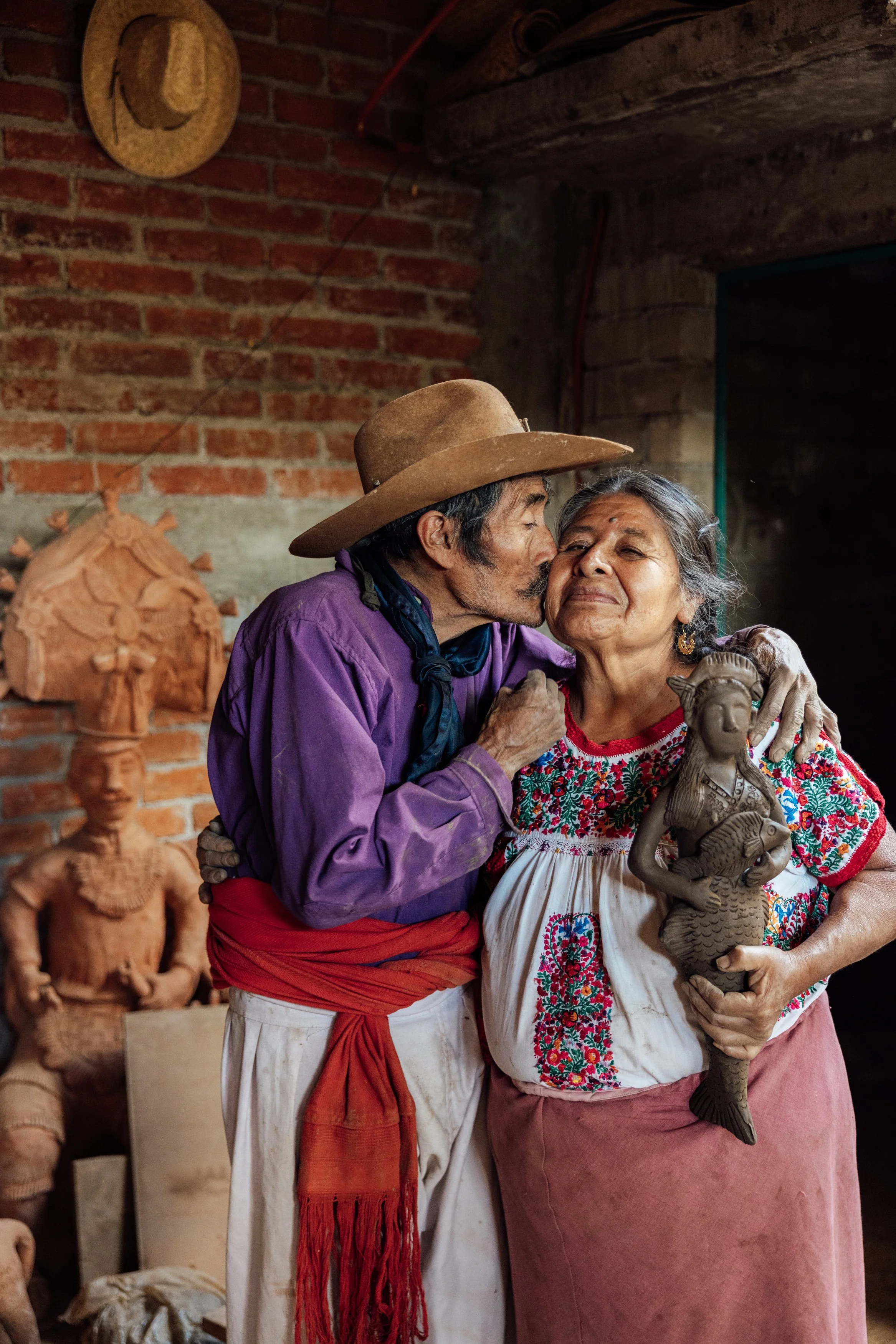 An elderly woman and a man sharing a kiss on the cheek inside a rustic workshop, with the woman holding a clay sculpture of a woman in traditional attire.