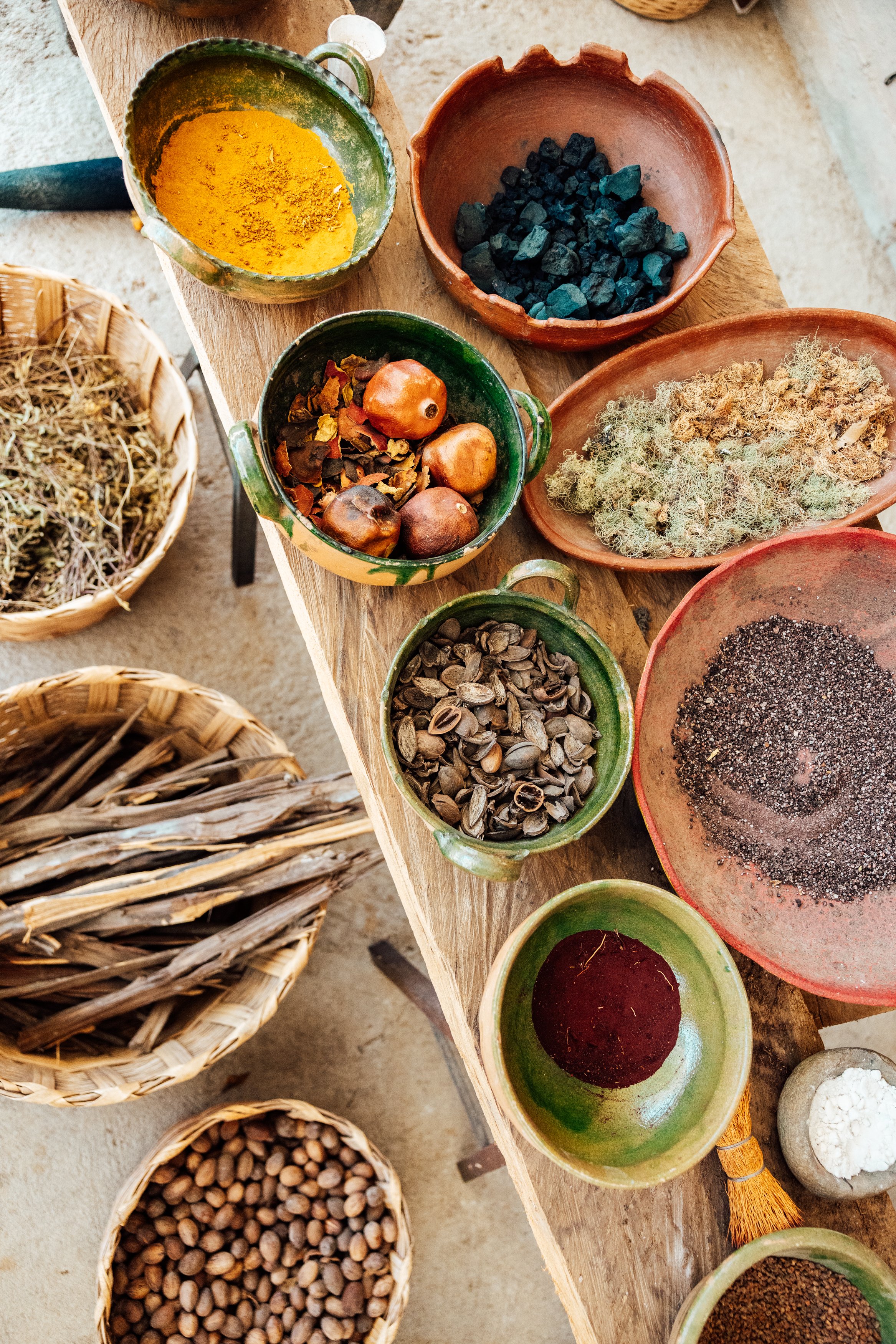 Various bowls and baskets of herbs, spices, and natural materials arranged on a wooden surface, including dried herbs, seeds, and colored powders, in a rustic setting.