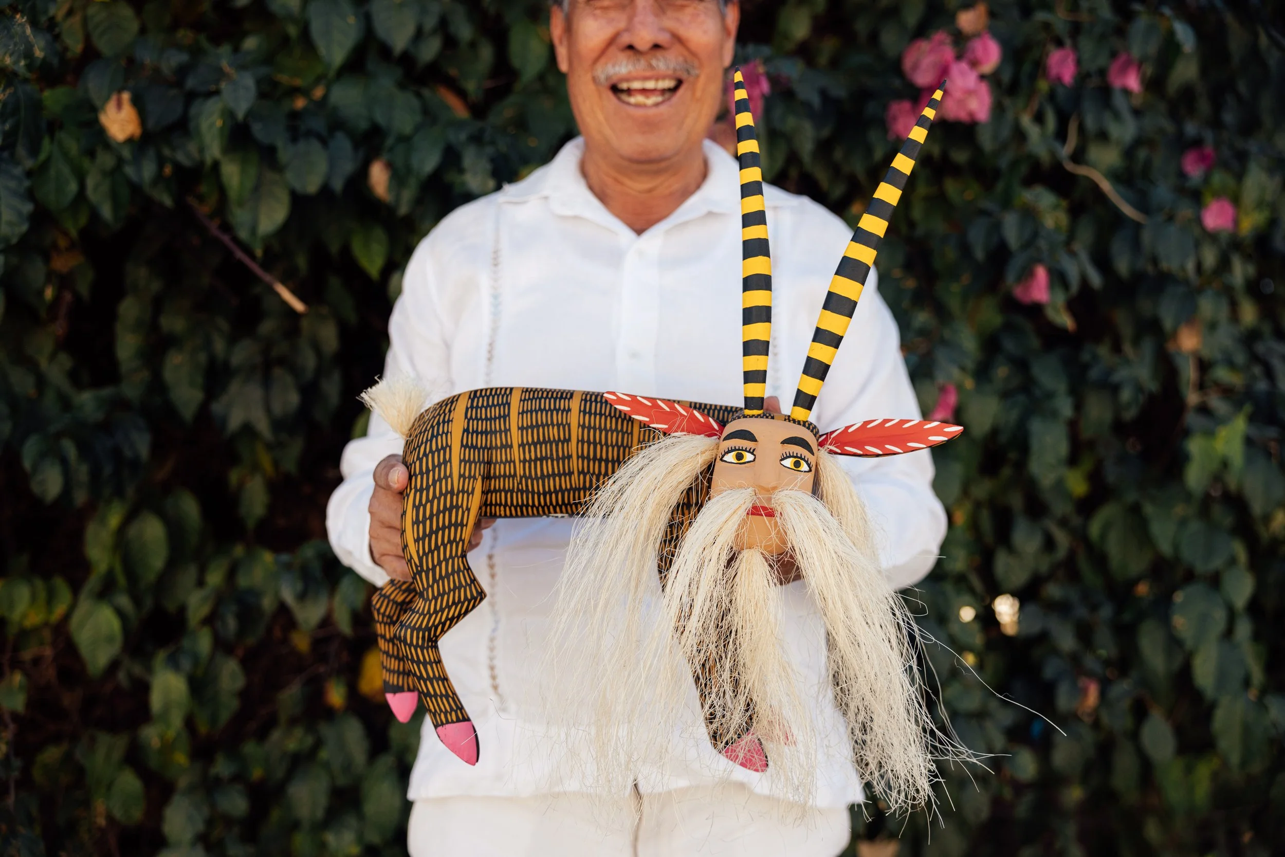 A man in a white shirt holding a colorful, decorative puppet of a character with long white beard, striped horns, and yellow eyes, outdoors with green foliage and pink flowers in the background.